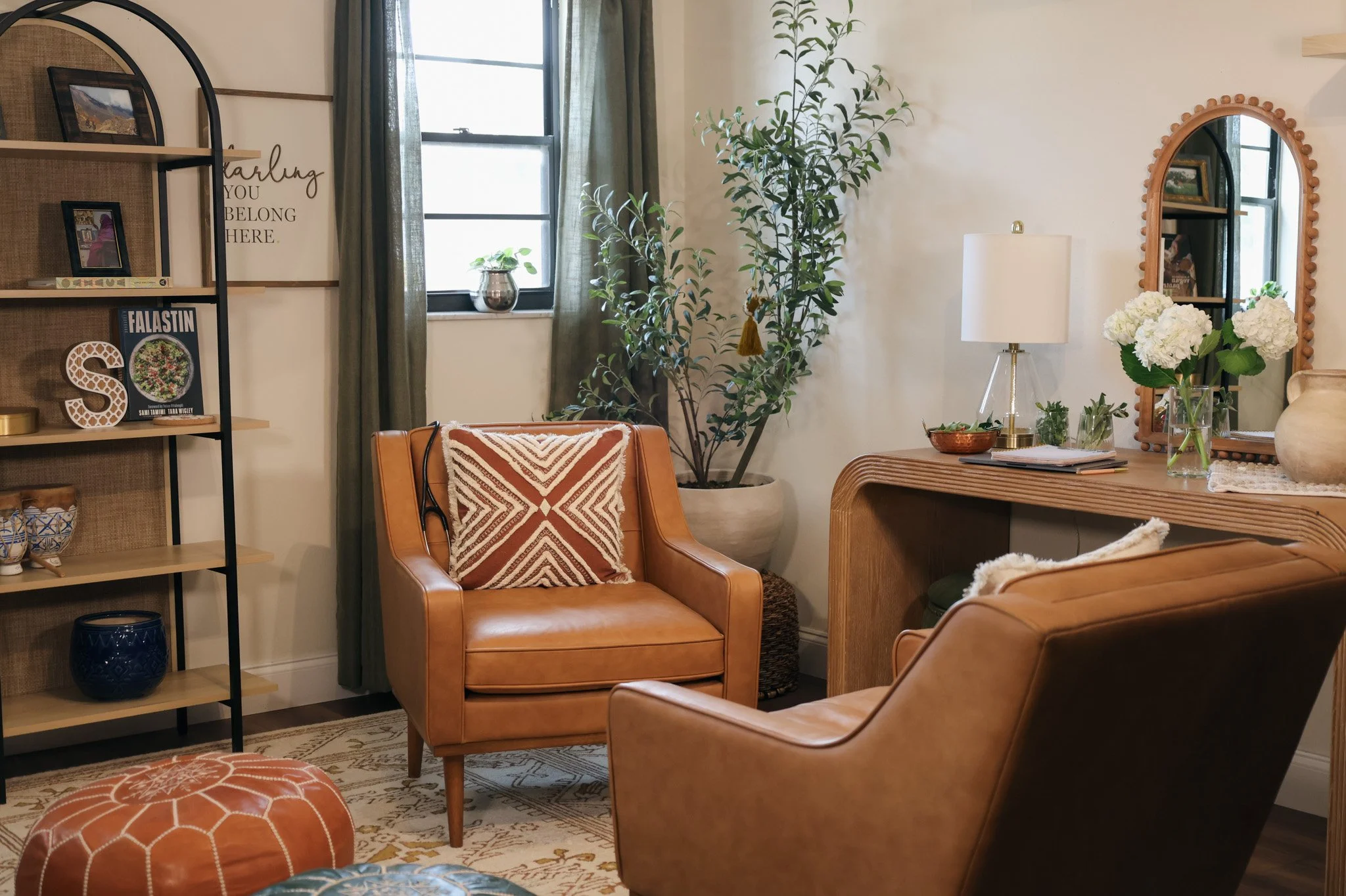 A cozy living room with tan leather chairs, a large potted plant, a framed mirror, a side table with a white lamp, and decorative items including flowers, framed pictures, and books. A window with curtains lets in natural light.