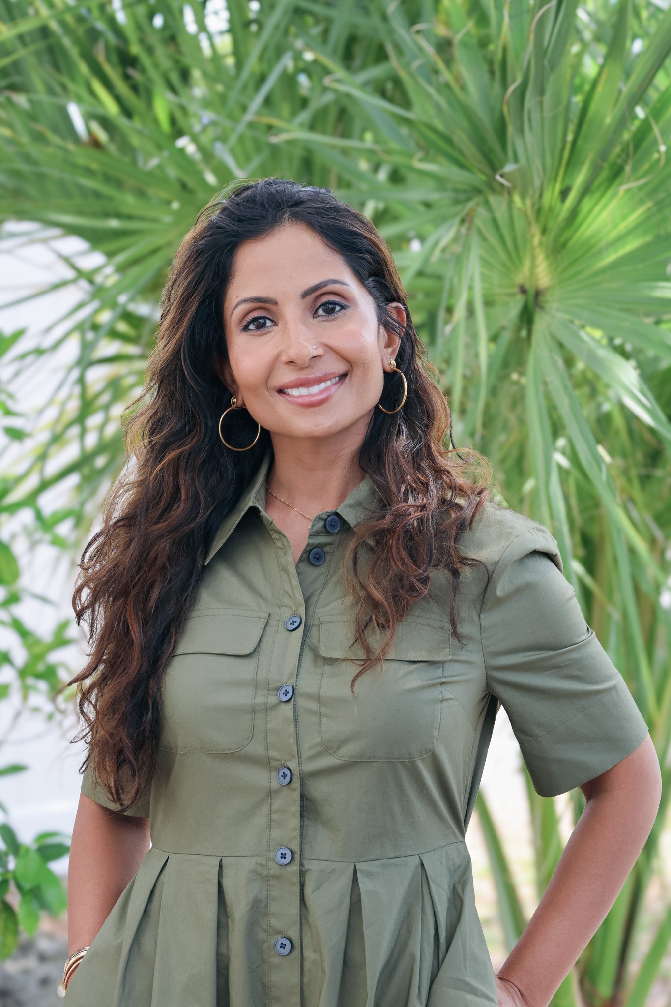 A woman with long dark hair, wearing gold hoop earrings and a green button-up dress, standing in front of green plants.