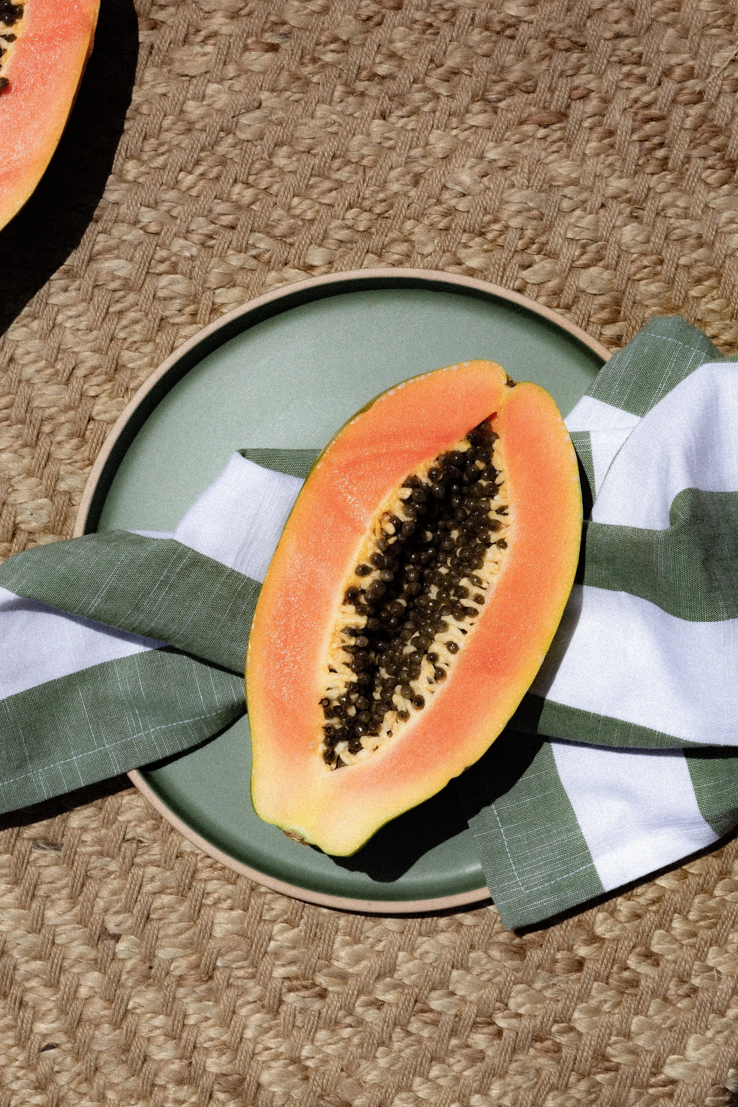 Half of a papaya fruit with orange flesh and black seeds on a green plate, placed on a green and white cloth napkin, on a woven surface.