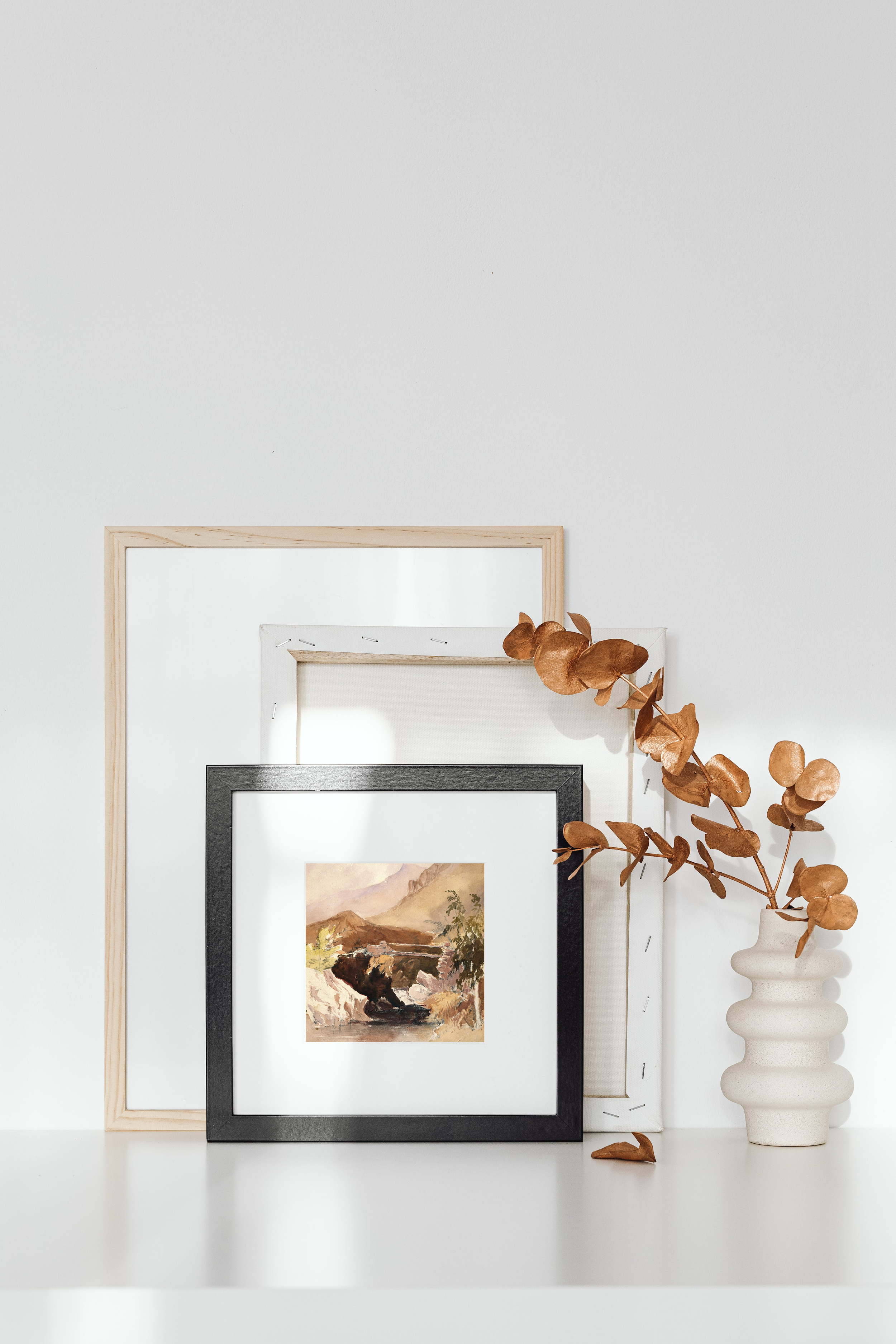 Arrangement of framed artwork leaning against a white wall, with dried leaves in a white ceramic vase beside them.