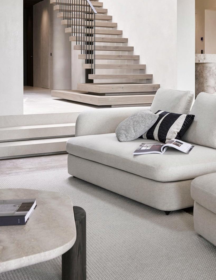 Living room with white sofa, black and white pillows, open magazine, marble coffee table, and a modern staircase with wooden steps and black metal railing.