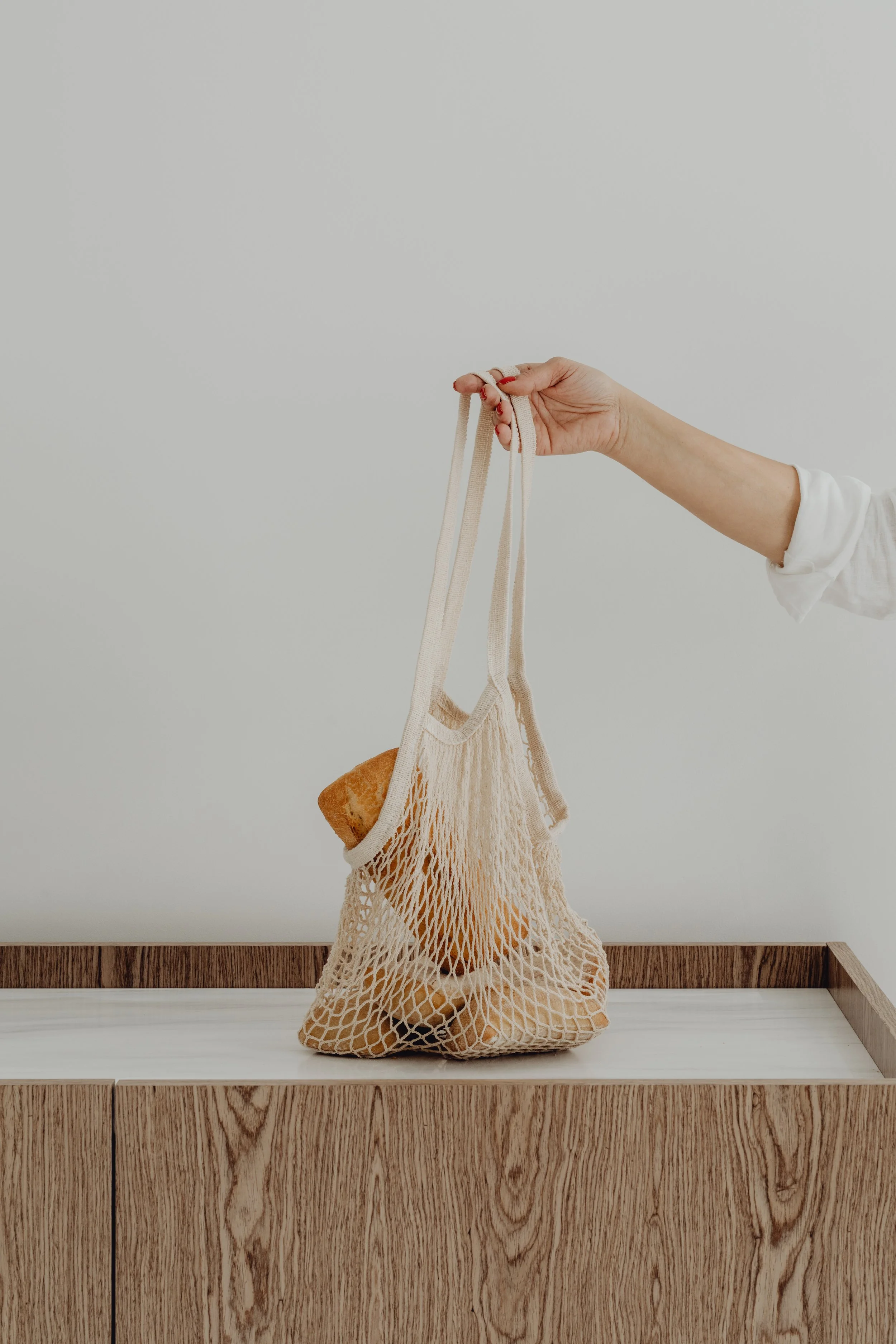 A woman holding beige reusable mesh bags with a loaf of bread inside, standing against a white wall with wooden paneling at the bottom.