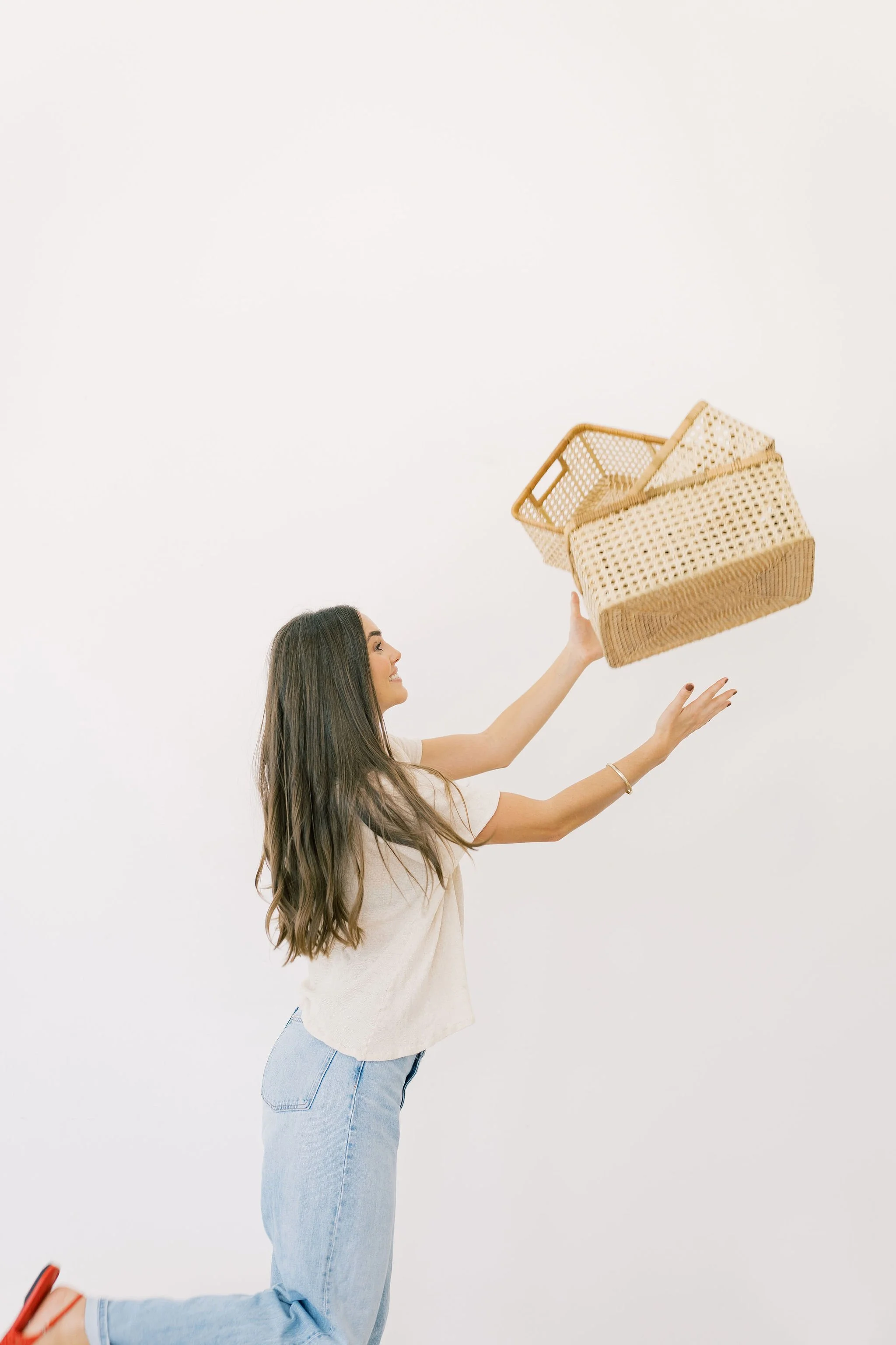 Woman with long brown hair kneeling and holding a wicker basket while smiling, against a plain white background holding woven organizing baskets