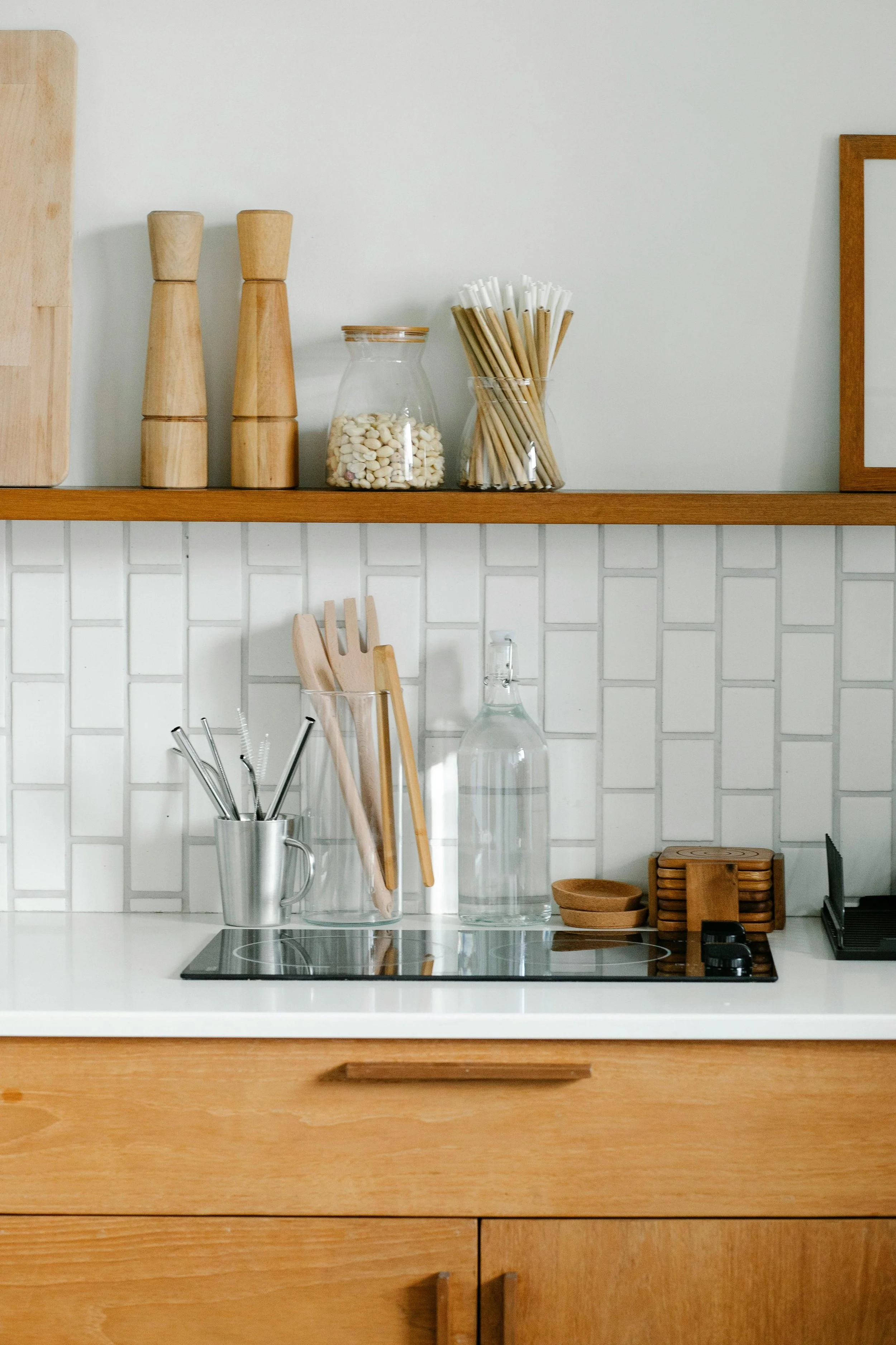 An organized kitchen counter with wooden cabinets, a stovetop, glass jar with white beans, utensils in a metal holder, glass water bottle, and small wooden bowls and coasters, with a wooden shelf above.