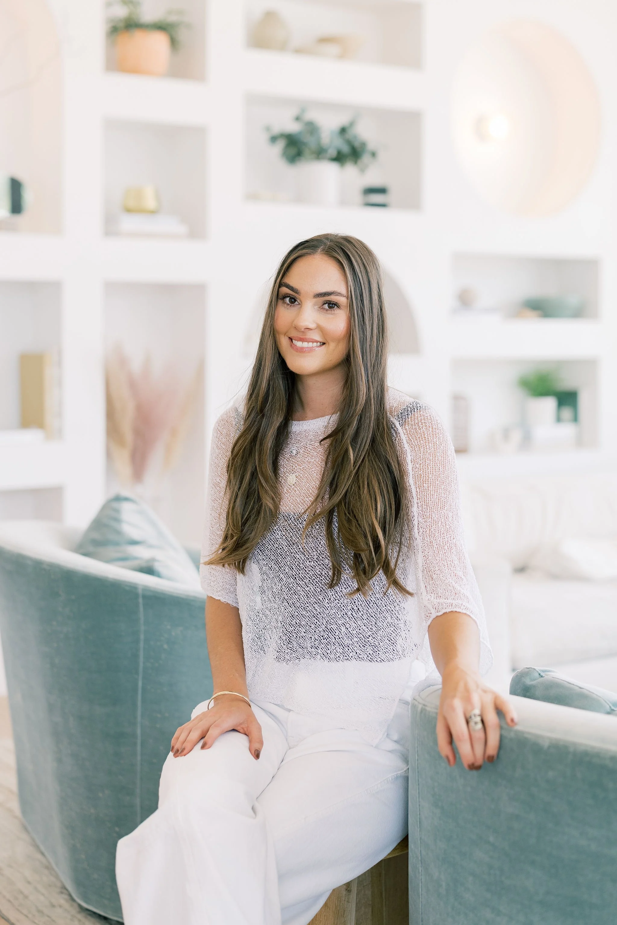 A young professional organizer woman with long brown hair, wearing a light pink knitted sweater and white pants, sitting on a teal-colored sofa in a bright, modern living room with white shelves and decorative items.