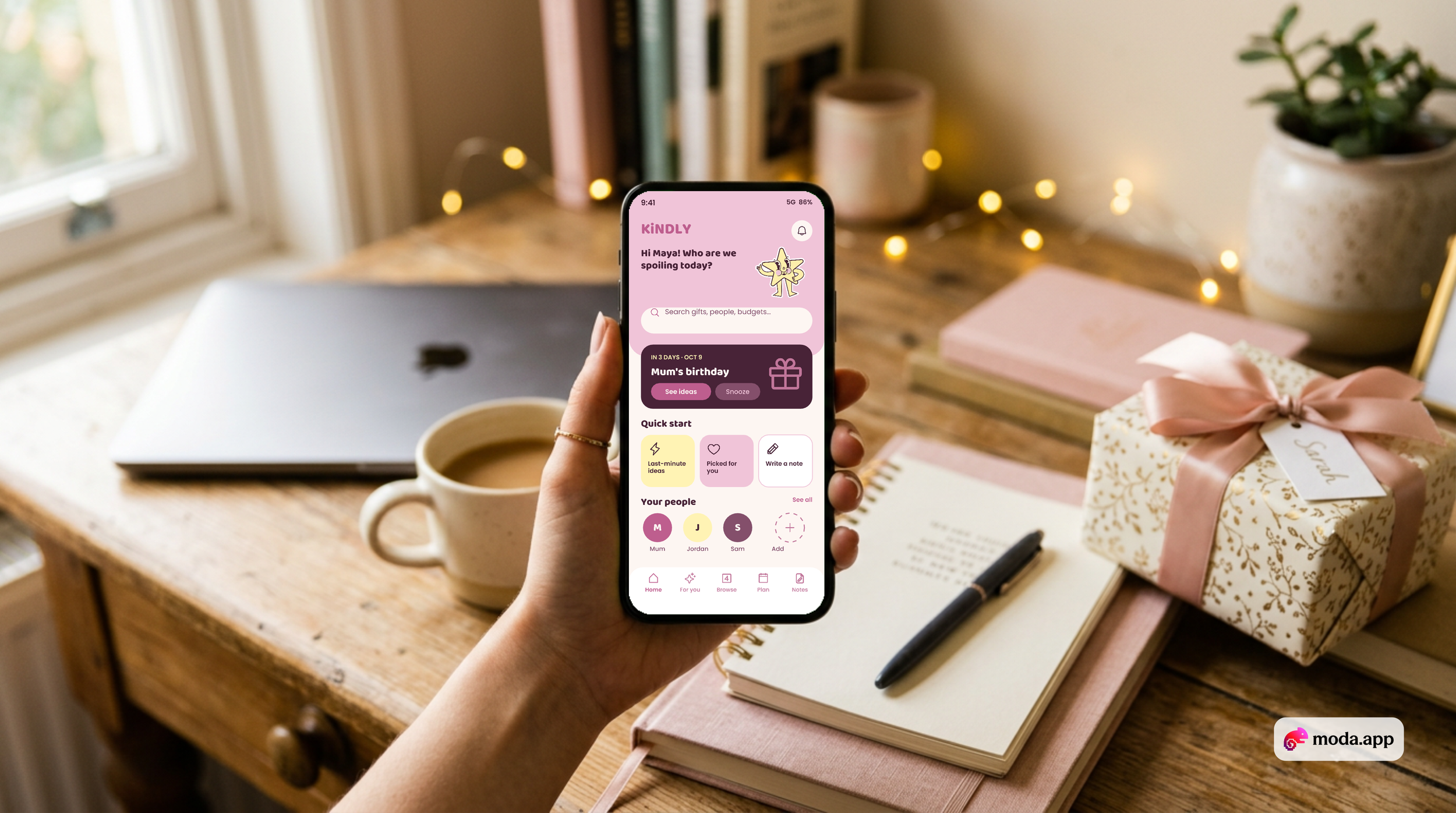 A person holding a smartphone displaying a gift planning app. The background shows a wooden table with a silver laptop, a cup of coffee, a pink notebook, a black pen, and a wrapped gift with a pink ribbon, near a window with sunlight and some decorative fairy lights.