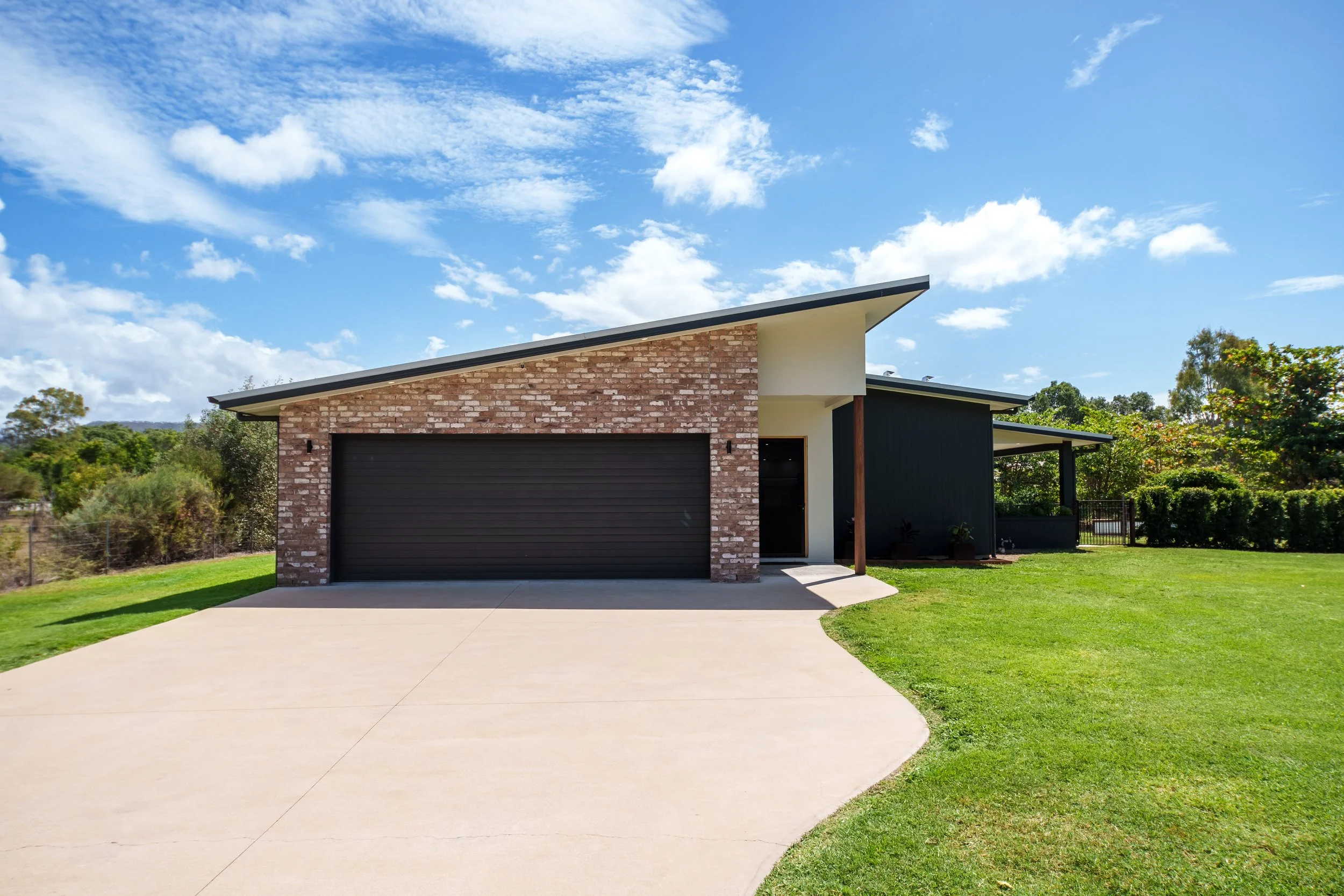 Modern house with brick and dark siding exterior, black garage door, and a sloped roof under a partly cloudy sky, surrounded by green lawn and trees.