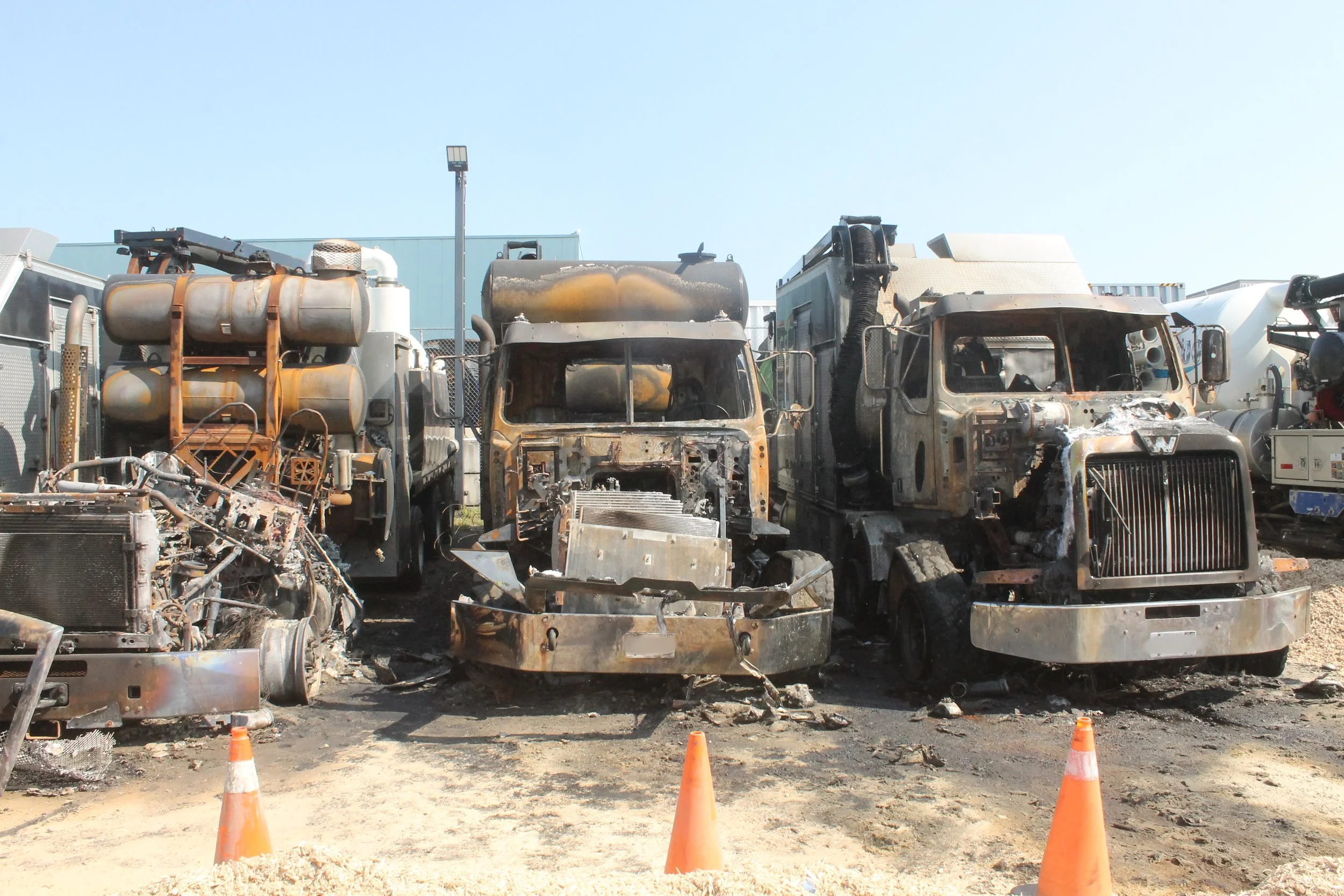 Burned and damaged trucks at a scrap yard with orange traffic cones in the foreground and a blue building in the background.