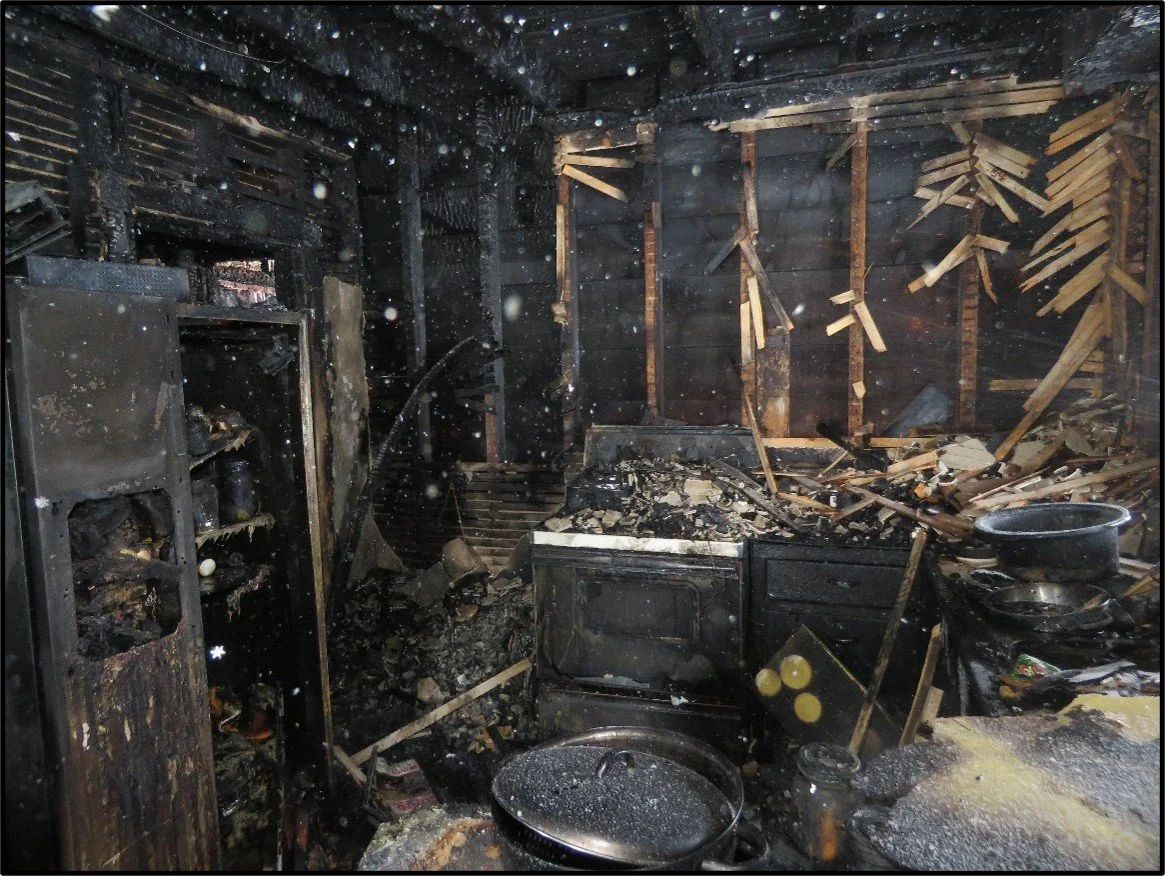A burned and charred kitchen with blackened walls, stove, cabinets, and burnt cookware scattered on the counter, after a fire.