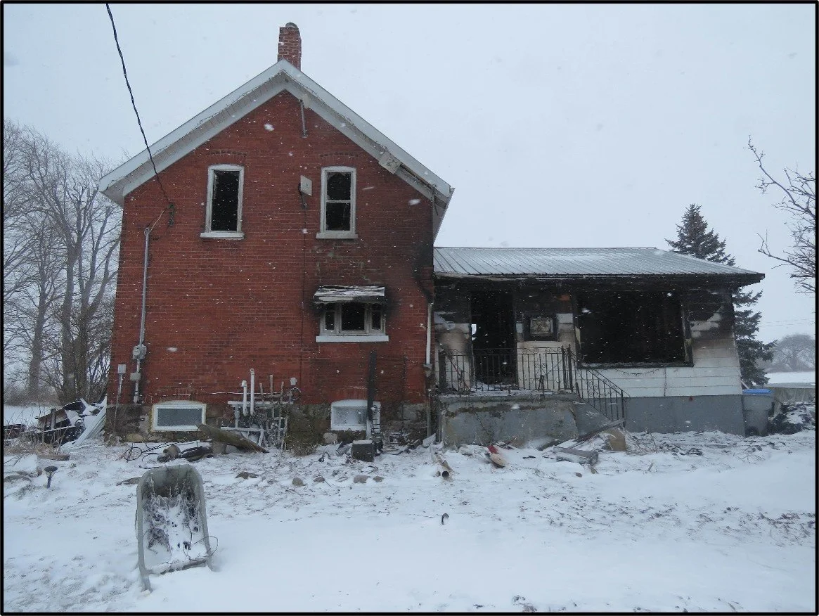 Burned and damaged house with broken windows, snow on the ground, and a snow-covered outdoor light fixture.