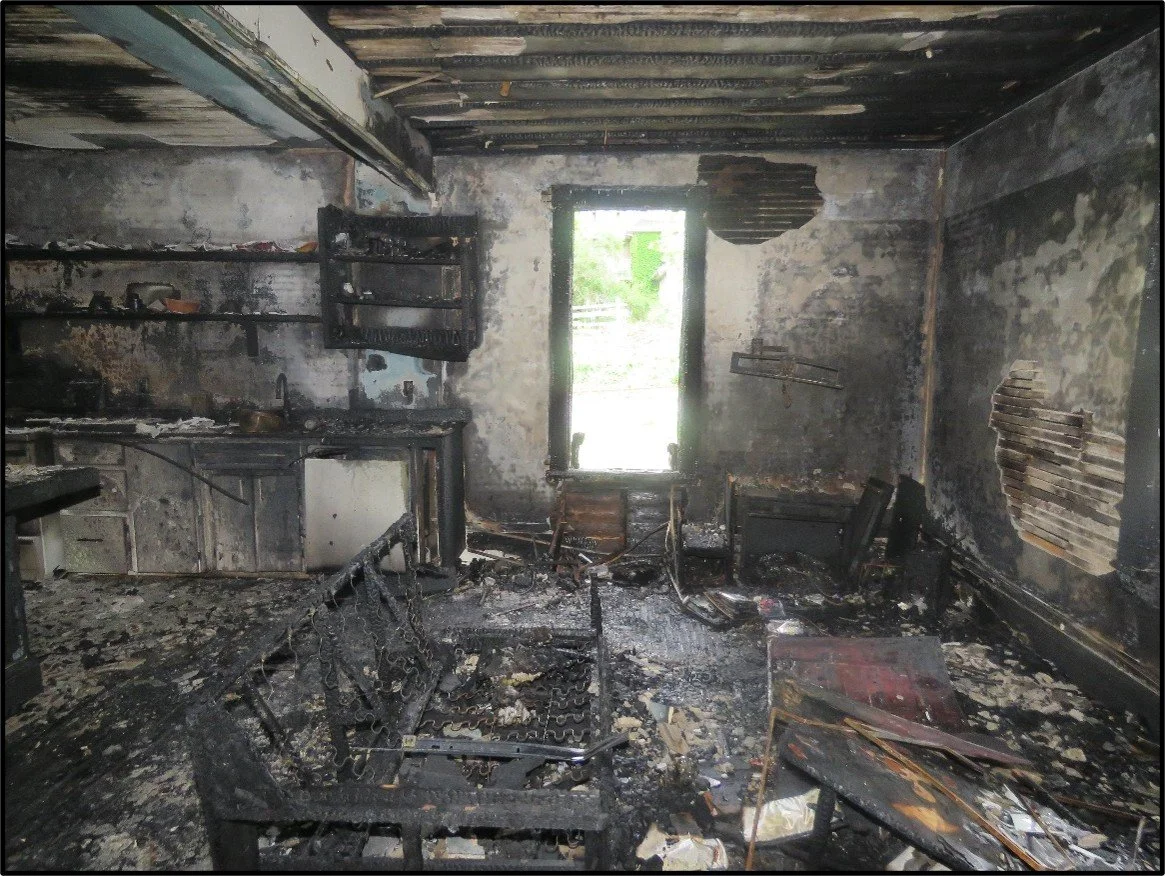 Room with blackened, charred walls and ceiling after a fire, with burnt furniture, debris, and a window providing bright light.