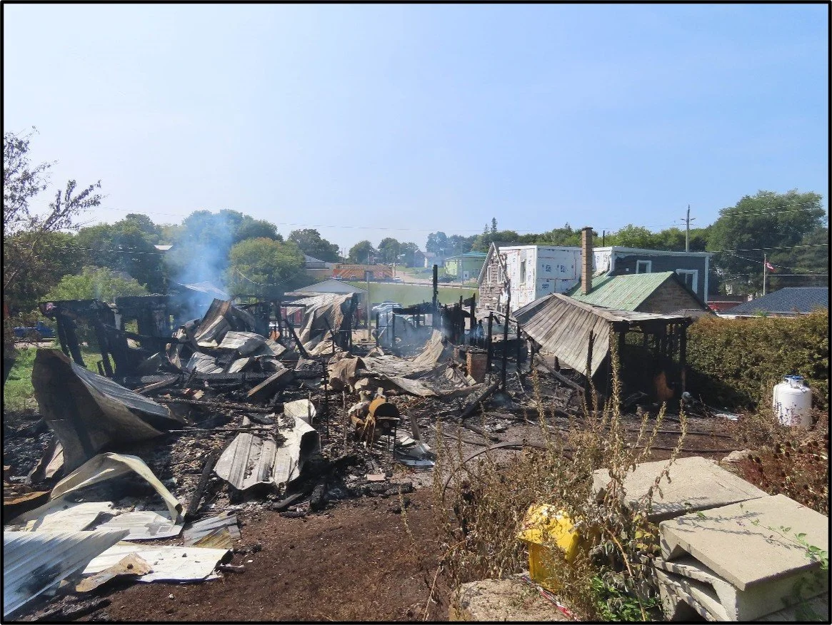 Burned and charred remains of a house or building after a fire, with smoke still rising, in a residential area.