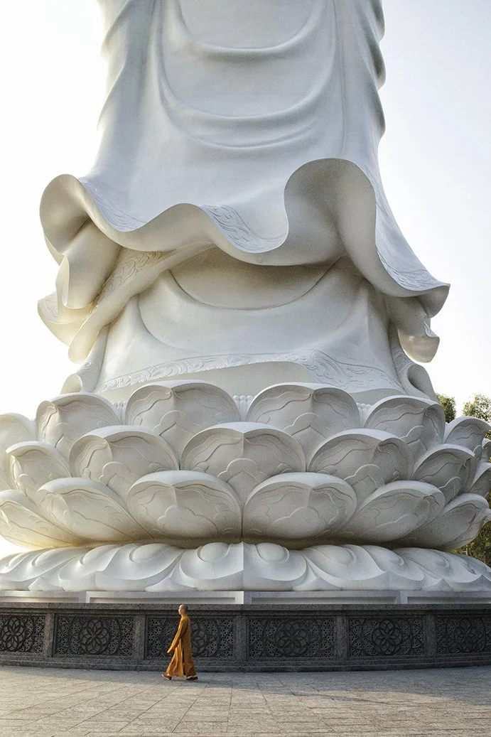 A large white statue of a religious figure, likely Buddha, situated on a lotus flower pedestal. A monk in traditional robes walks in front of the statue.