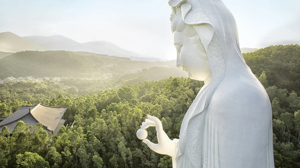 A large white statue of a woman holding a sphere in her hand, overlooking a lush green landscape with trees and mountains in the background.