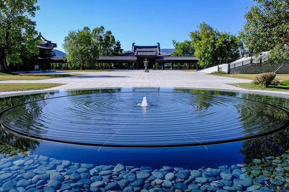 A circular water fountain with ripples in the foreground, surrounded by rocks. In the background, there are traditional East Asian-style buildings with curved rooftops, trees, and a clear blue sky.