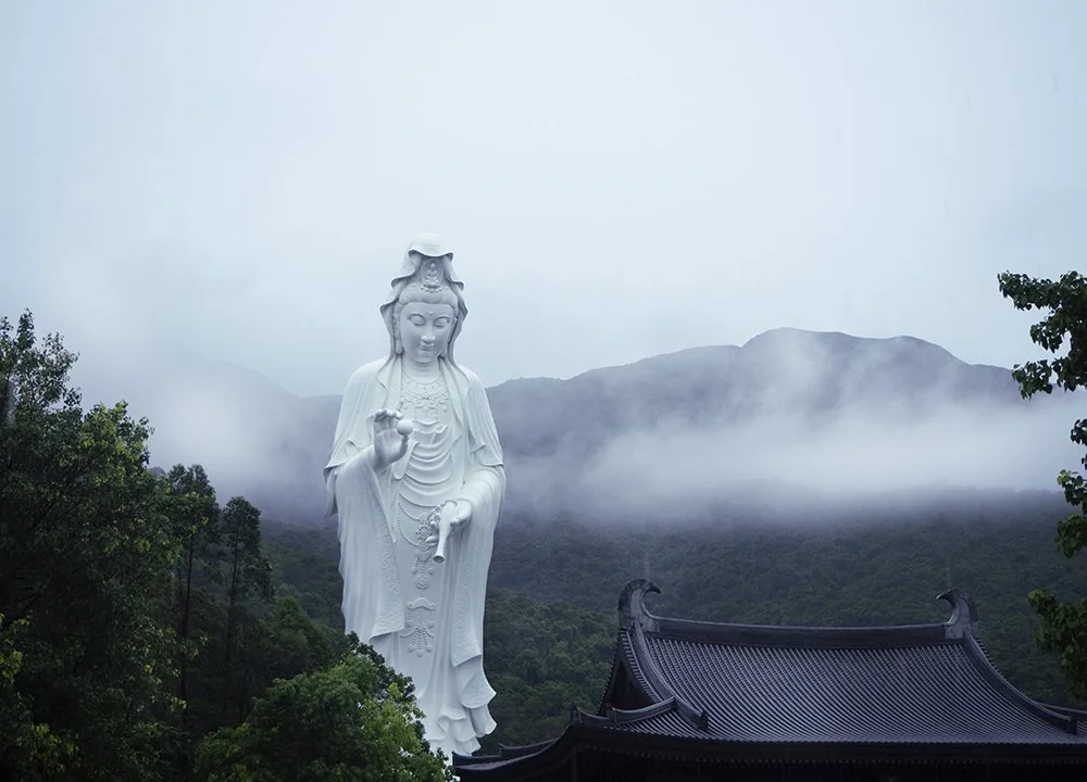 A large white statue of a Buddhist figure, likely a Bodhisattva, stands in a mountainous landscape with fog and greenery. A traditional dark-colored temple roof is visible at the bottom.