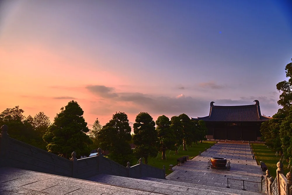 A traditional Asian temple at sunset with a cloudy sky, surrounded by trees and stone steps leading to the temple entrance.
