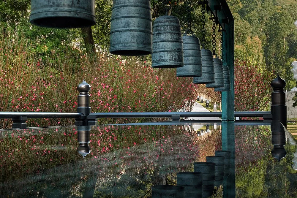 A row of large, old-fashioned hanging bells suspended from a curved green metal frame over a reflective surface. Behind the bells, there are flowering bushes with pink blossoms and tall trees with green foliage.