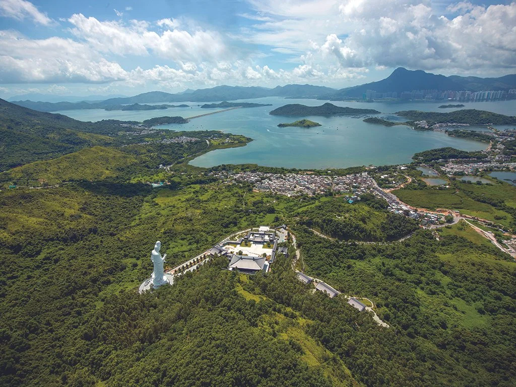 Aerial view of a large, white statue of a deity overlooking lush green hills, a river, and a small town with mountains in the background.