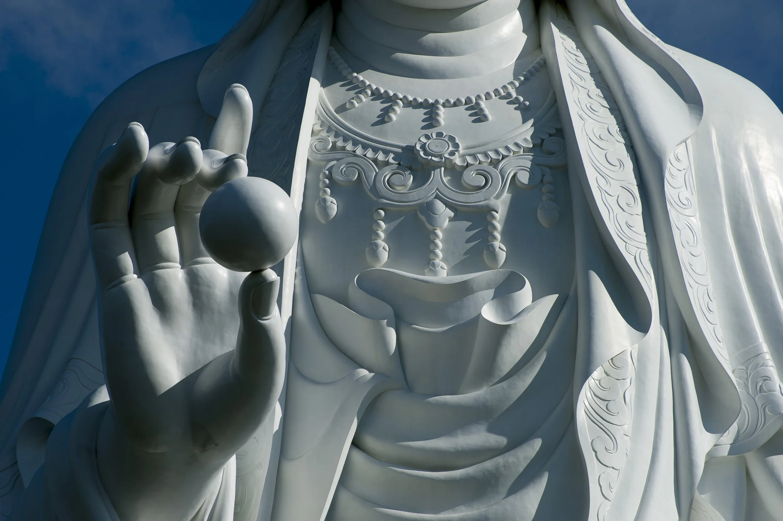 Close-up photo of the intricate carved details on the large white statue of a seated figure, with one hand holding a round object and wearing multiple necklaces and robes, against a bright blue sky.