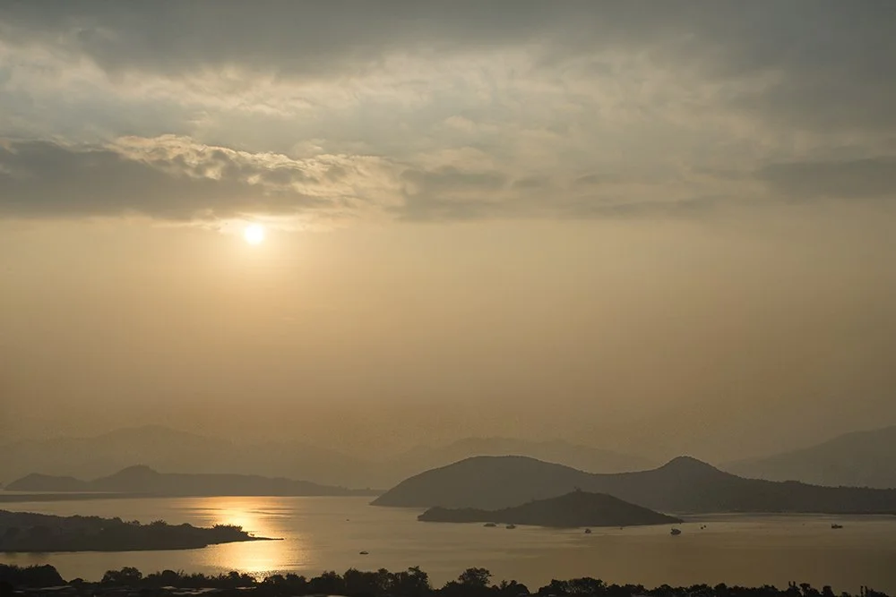 Sun setting over a lake surrounded by mountains with cloudy sky.
