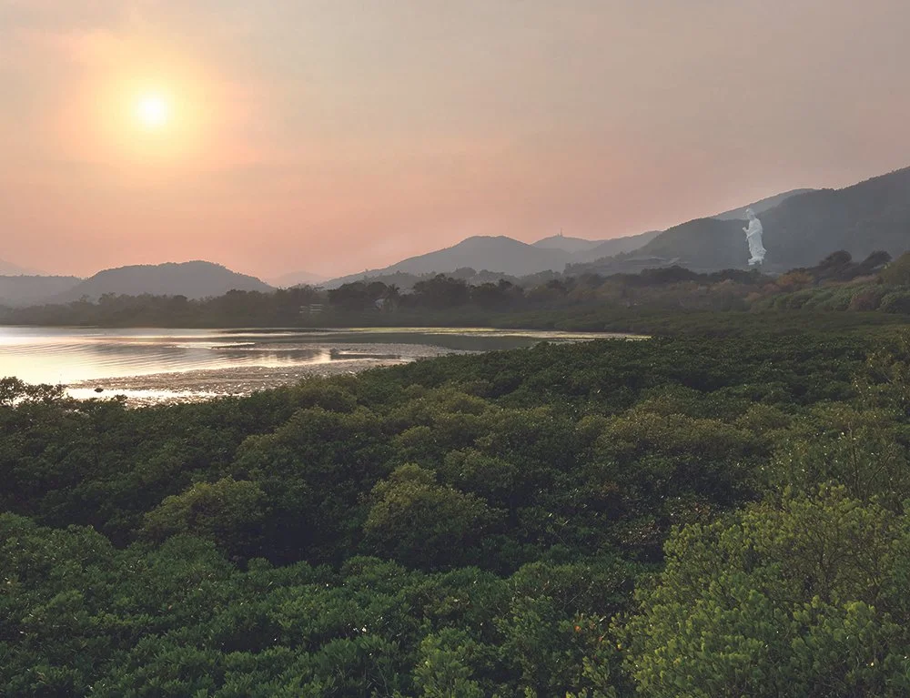 Sunset over a body of water surrounded by lush green foliage and distant mountains with a smokestack emitting smoke in the background.
