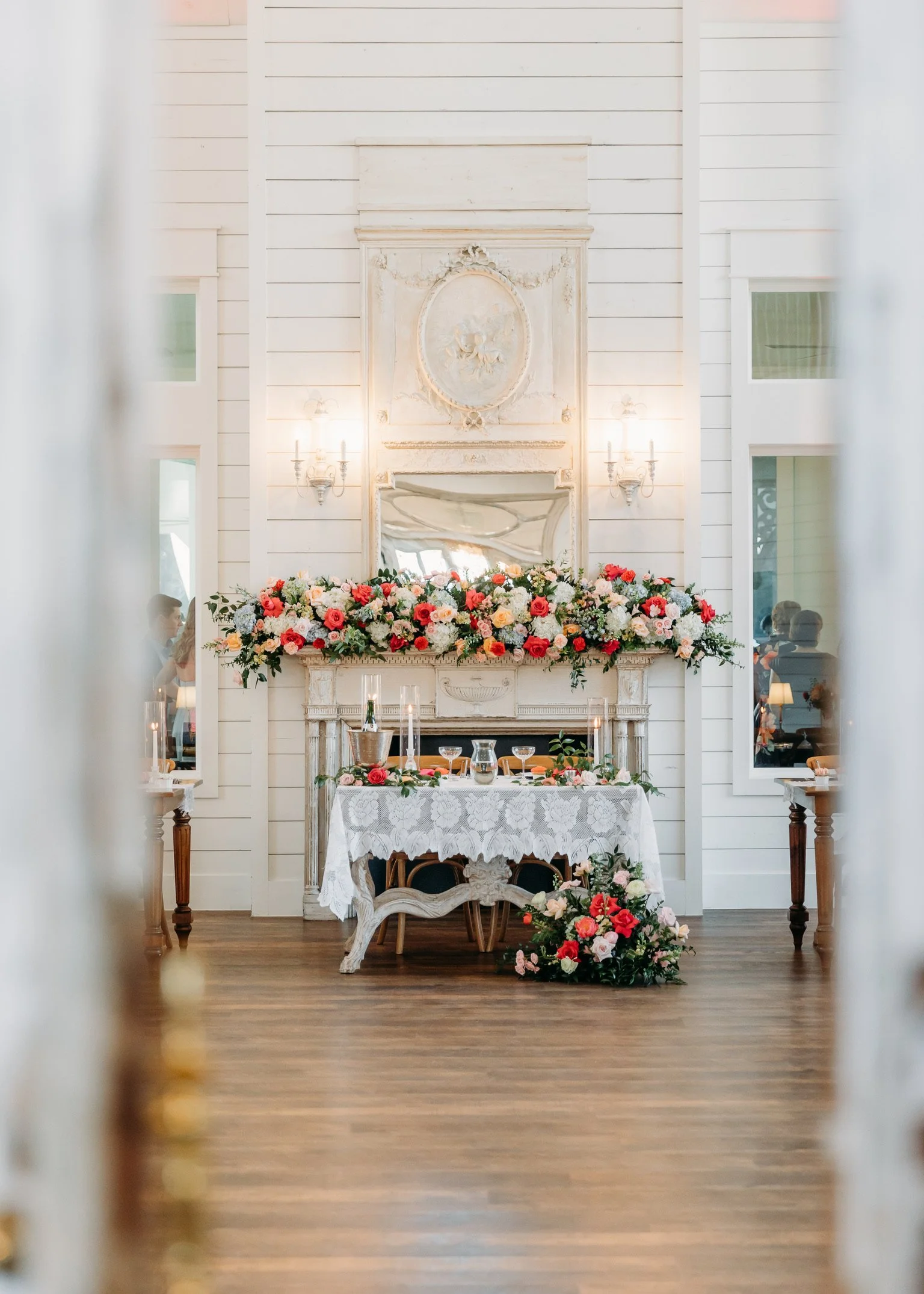 Wedding reception setup with a table covered by a white lace tablecloth, decorated with floral arrangements, candles, and glassware. Behind the table, there is a fireplace with a floral garland and an ornate mirror. The room has white walls with deco