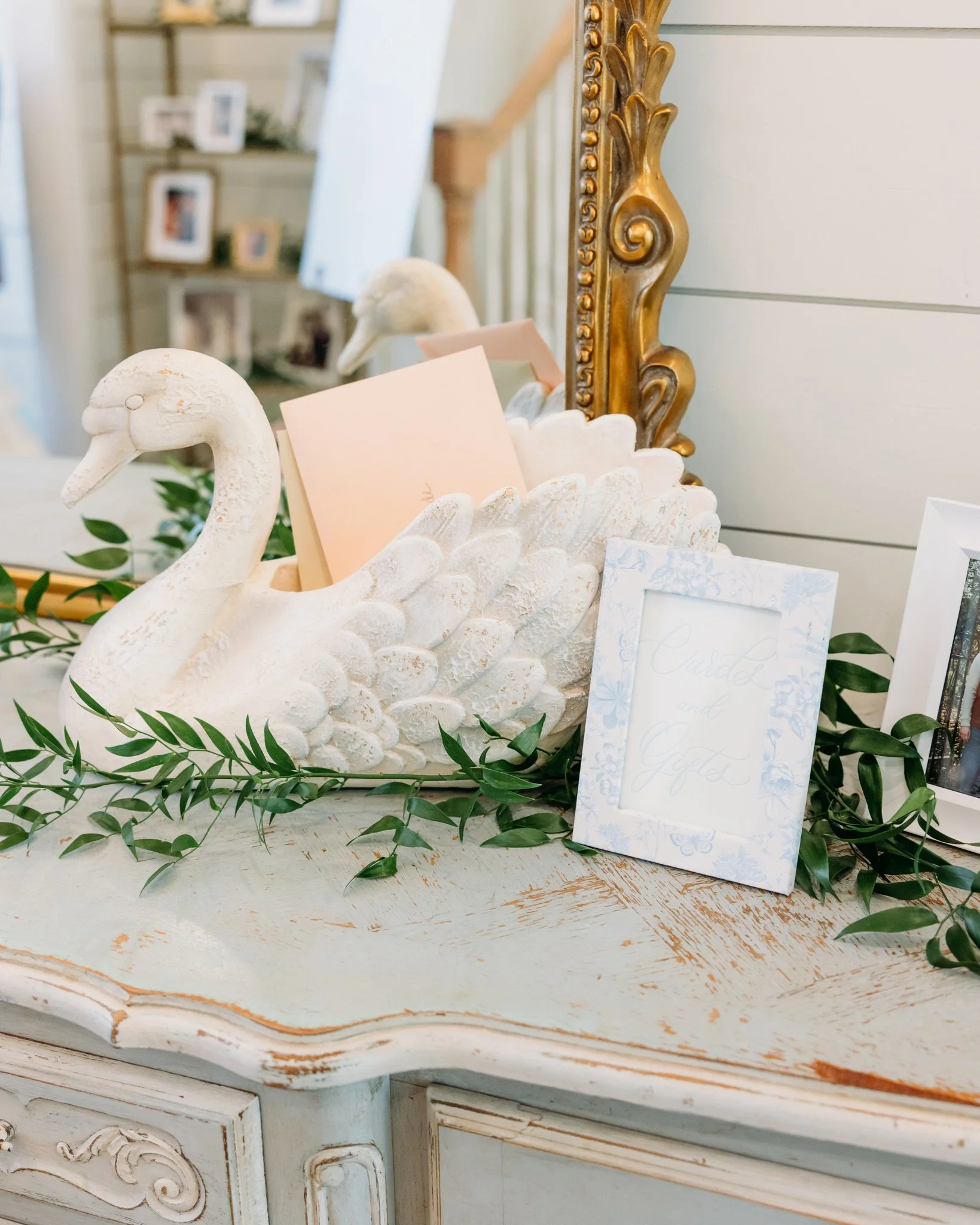 An ornate, vintage mirror with a gold, decorative frame leaning against a wall. In front of the mirror, there is a swan sculpture with a long neck, painted white with a weathered look. The sculpture has a few pastel-colored cards or notes placed on i