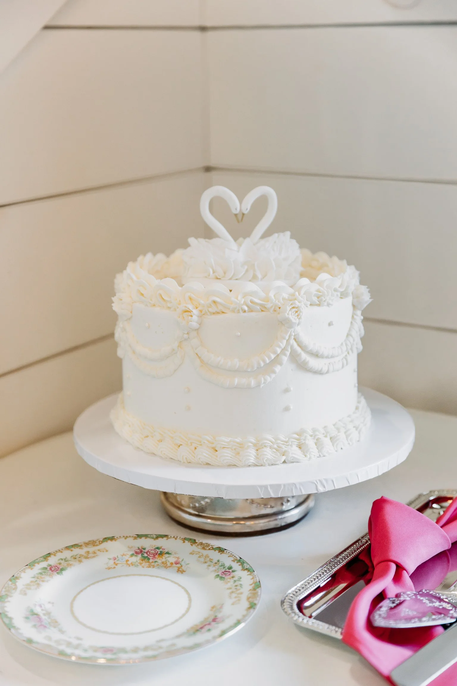 A white cake with piped icing and a heart-shaped topper, sitting on a cake stand, with decorative plates and pink napkins nearby.