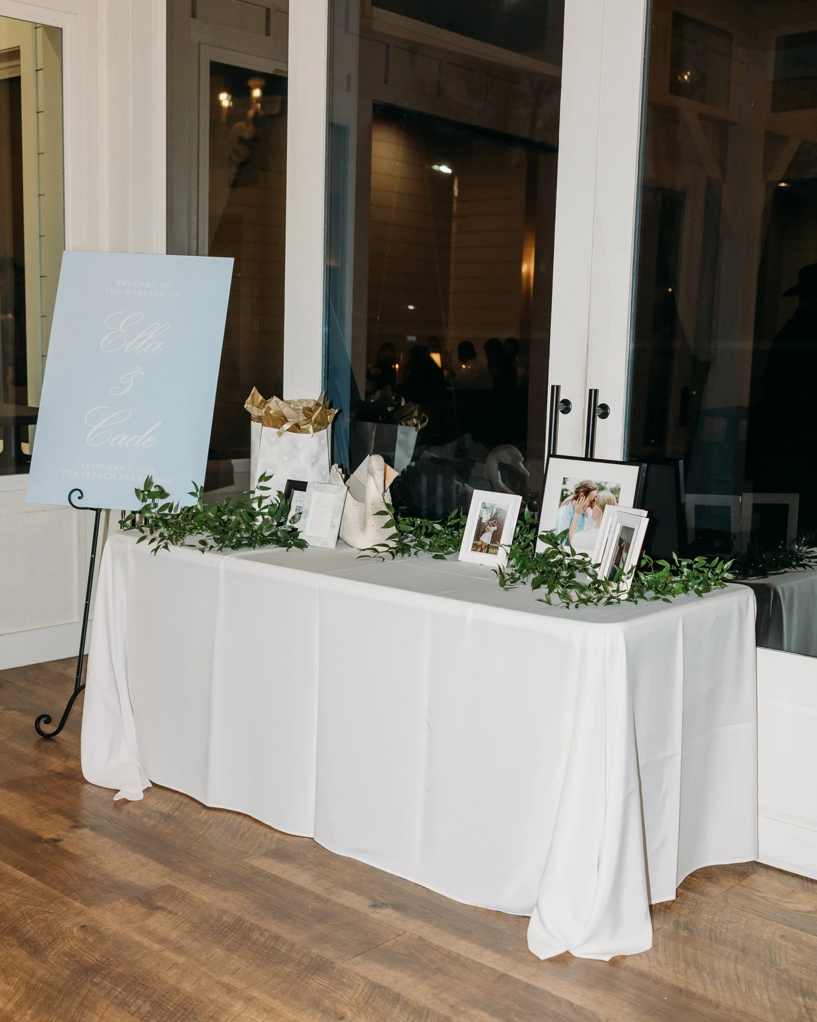 A wedding reception table decorated with framed photos, green foliage, and presents, with a blue wedding sign in the background sign welcoming guests to the wedding of Ella and Cade, February 7, 2023.