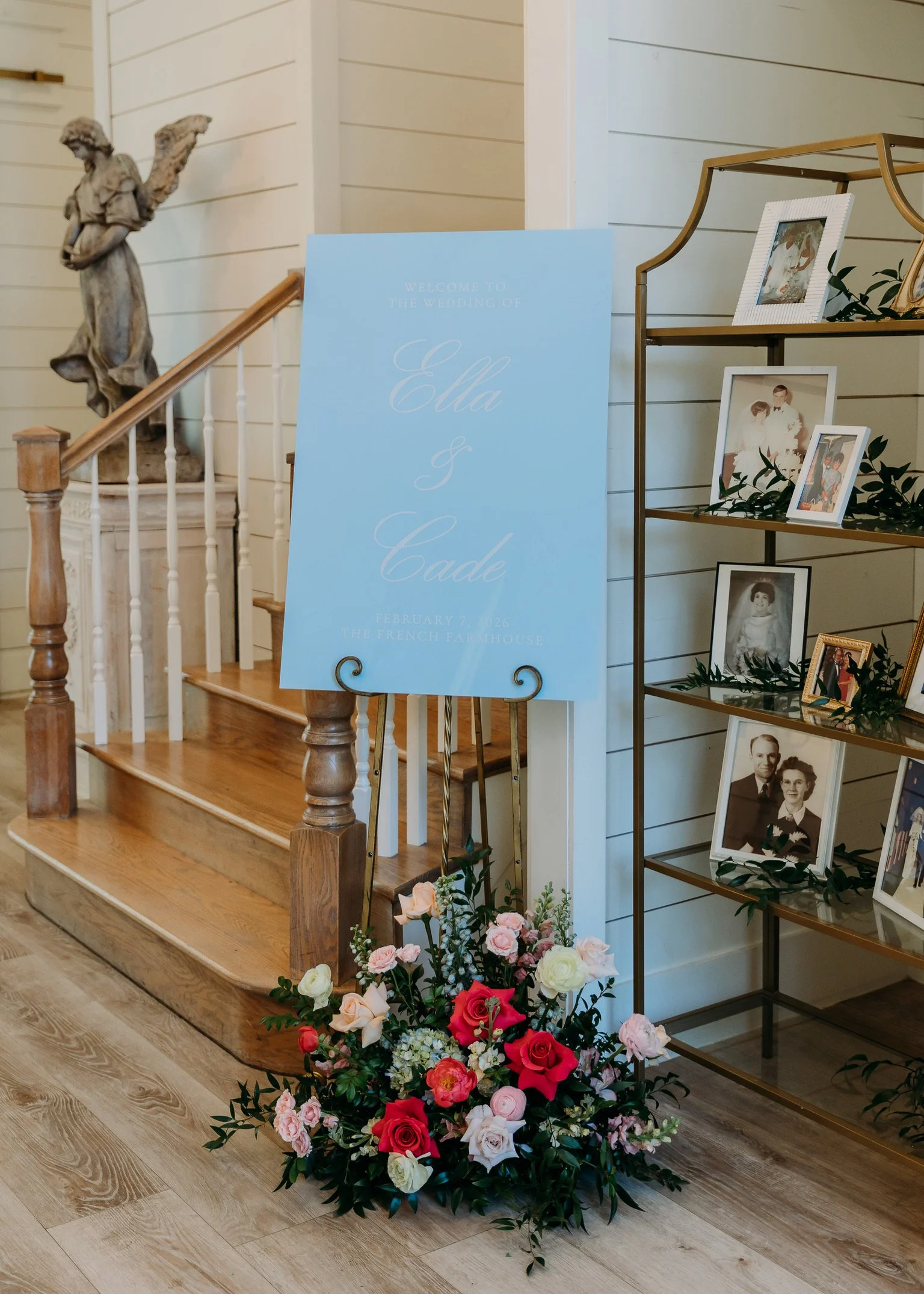 Flower arrangement with pink, white, and red roses along with greenery at the base of a wedding sign and a photo display shelf in a home or venue setting.