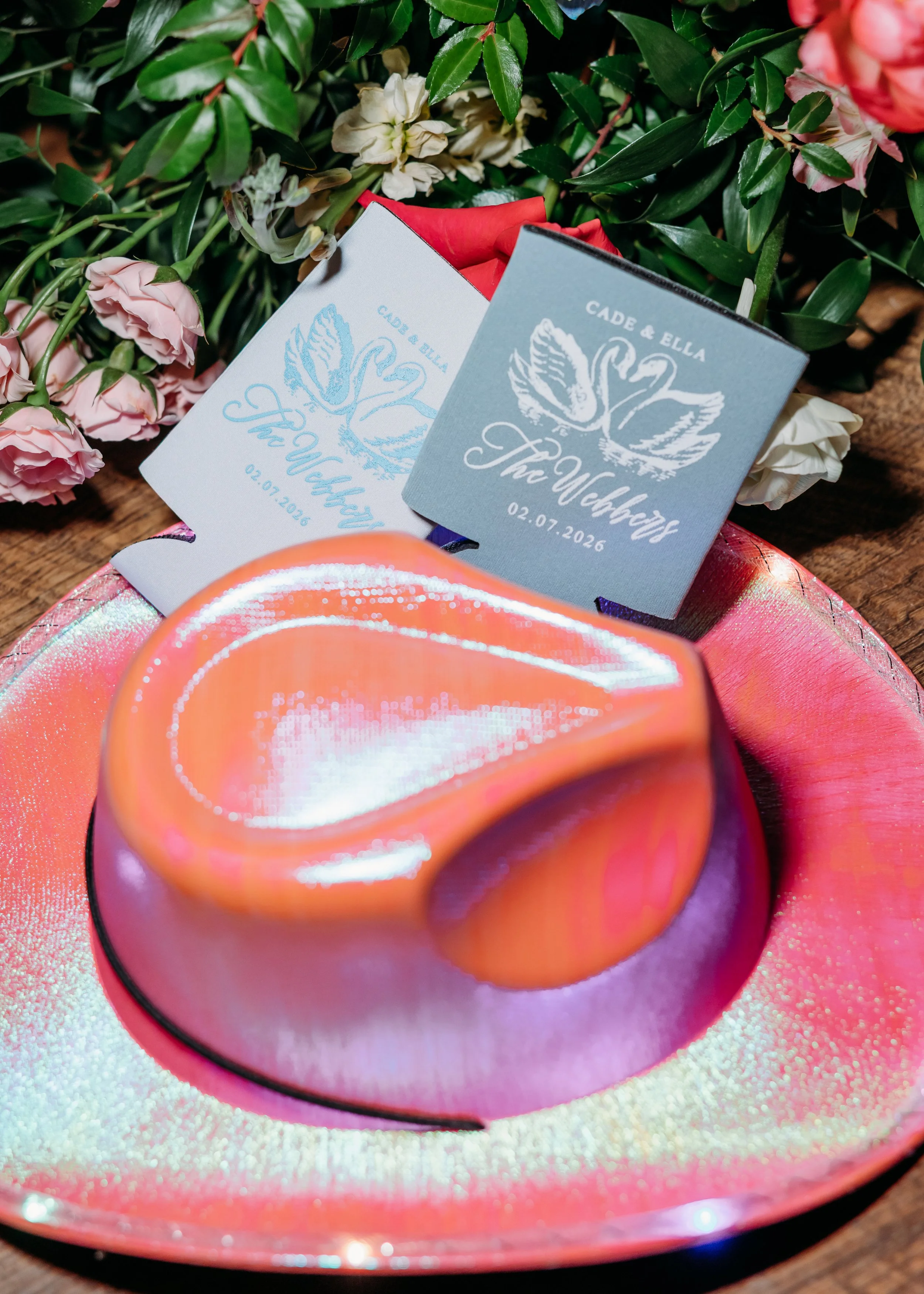 Close-up of two wedding program flyers with a swan and floral design, placed among pink and white flowers on a wooden surface. One flyer is light blue, and the other is white with blue text. The date 02.07.2026 and the names Cade & Ella are visible.