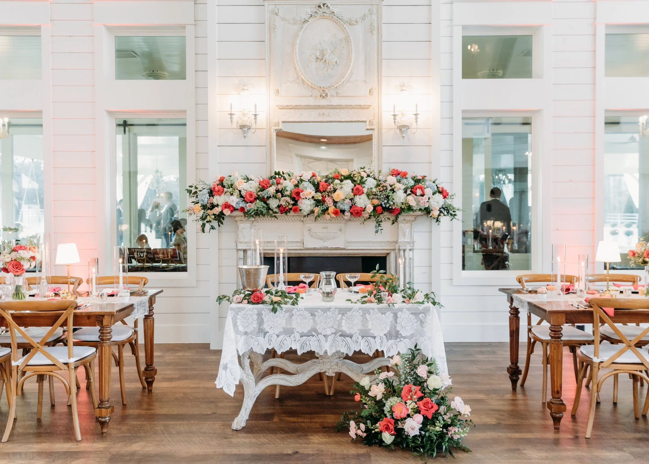 A decorated indoor space for a wedding or event, featuring a fireplace with a floral arrangement, a mirror, and wall sconces. Nearby tables are set with flowers and candles, surrounded by wooden chairs with white cushions.