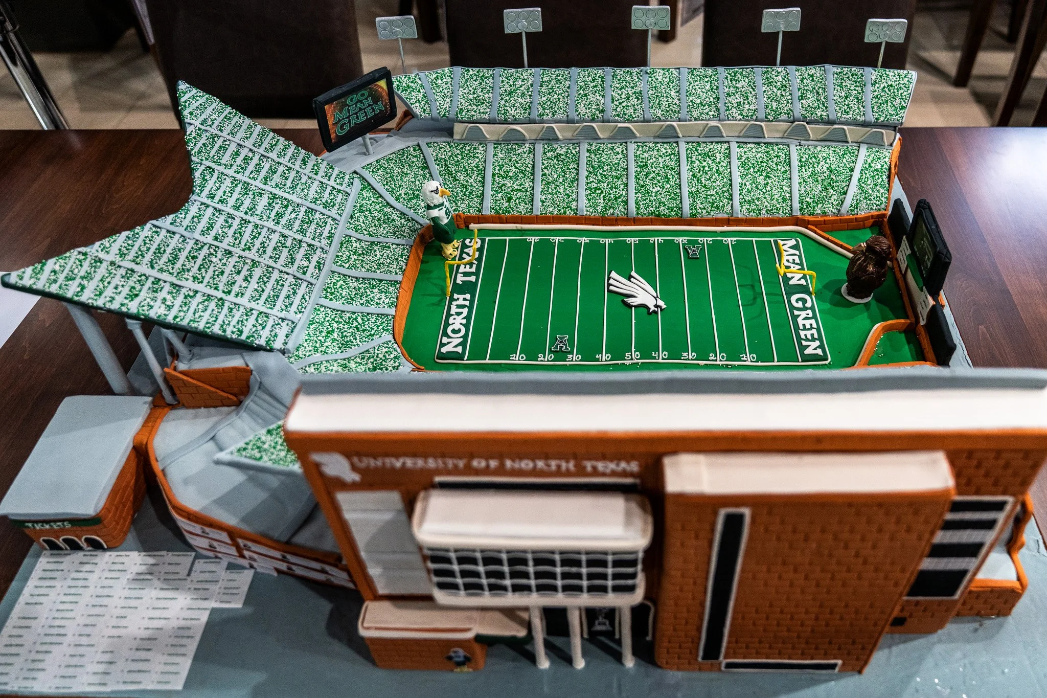 A model of a football stadium with a green field, showing the University of North Texas logo and the words 'Go Mean Green' on a screen, with a mascot statue and seating sections made of foam and plastic.