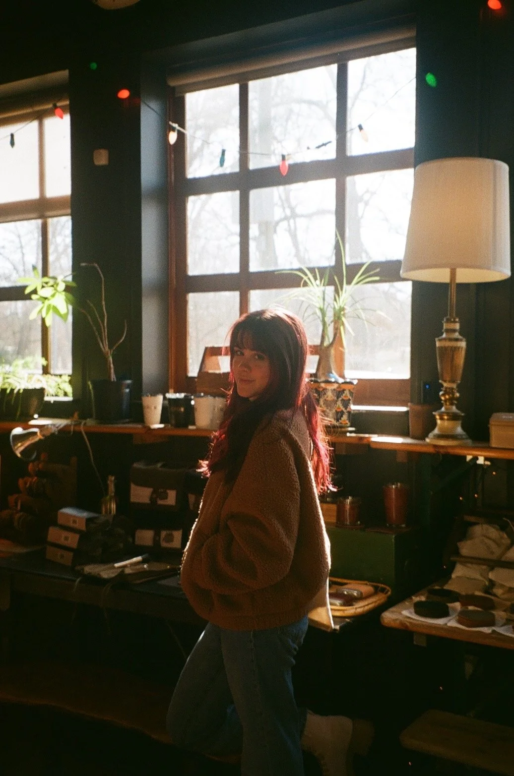 A woman with dark hair and a brown jacket stands inside a room with large windows, plants, a table, and a lamp, with sunlight streaming in.