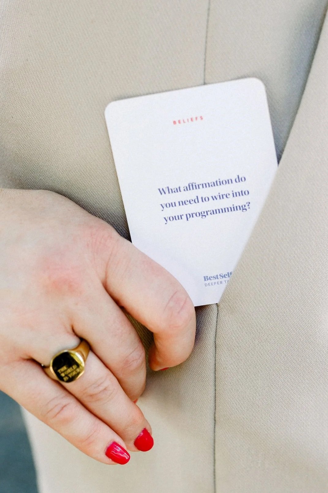 A person's hand with red nail polish and a gold ring holding a card that reads 'What affirmation do you need to wire into your programming?'' against a beige fabric background.