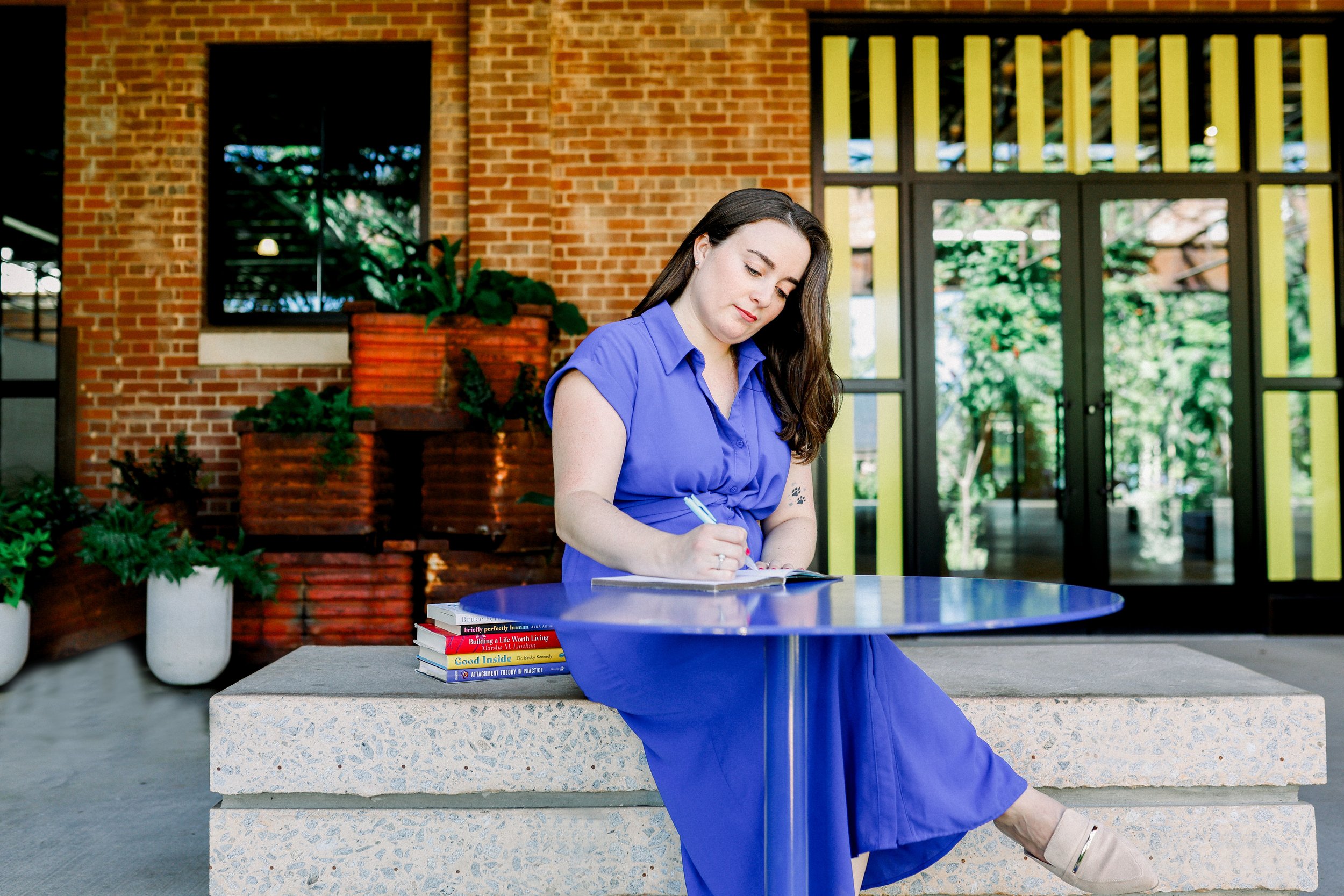 A woman in a blue dress sitting at a blue table and writing in a notebook, with a stack of books underneath the table, in an outdoor area with brick walls, windows, and plants.