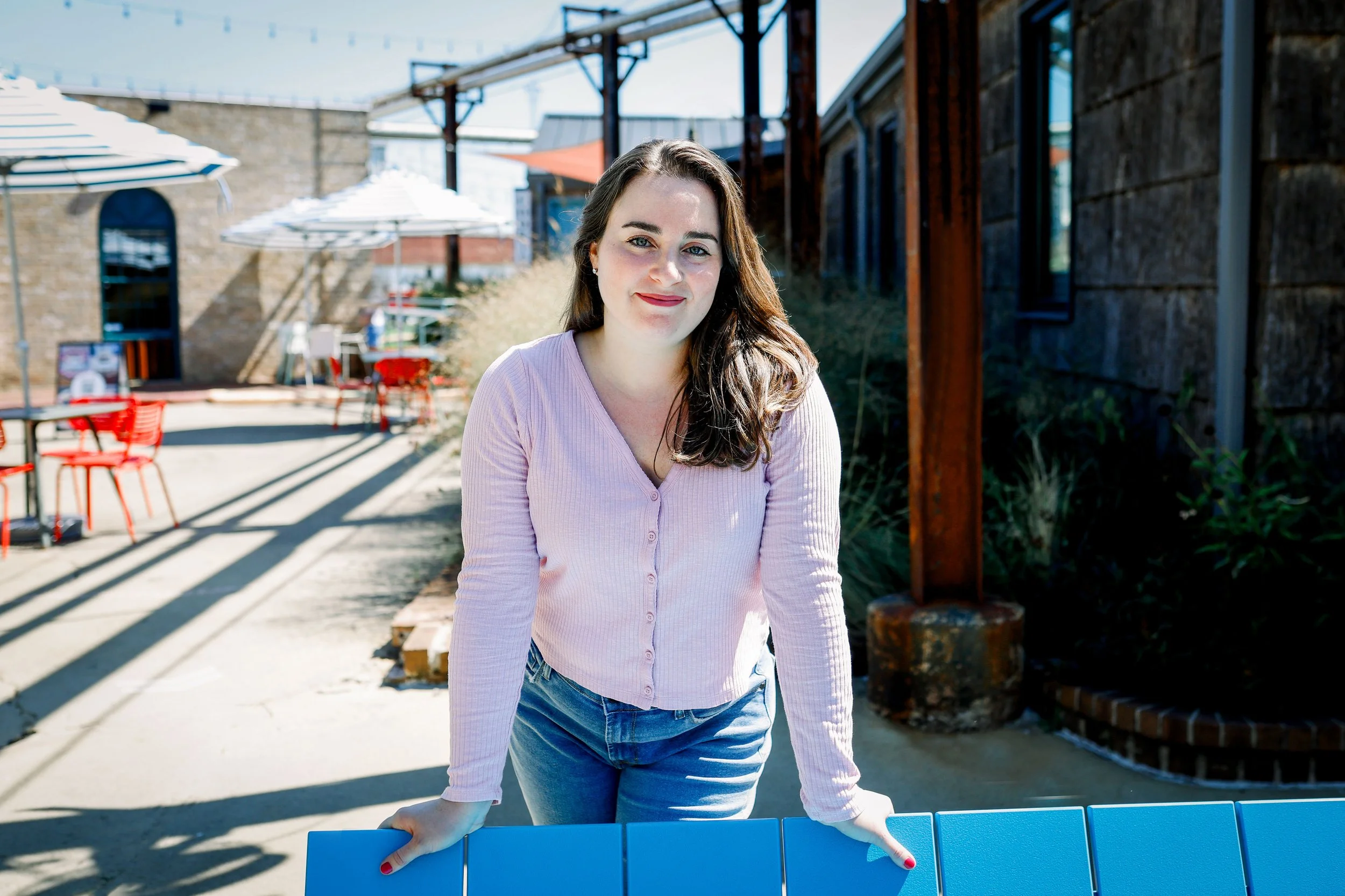 A woman with brown hair wearing a pink shirt and jeans, smiling outdoors on a sunny day with umbrellas and outdoor seating in the background.
