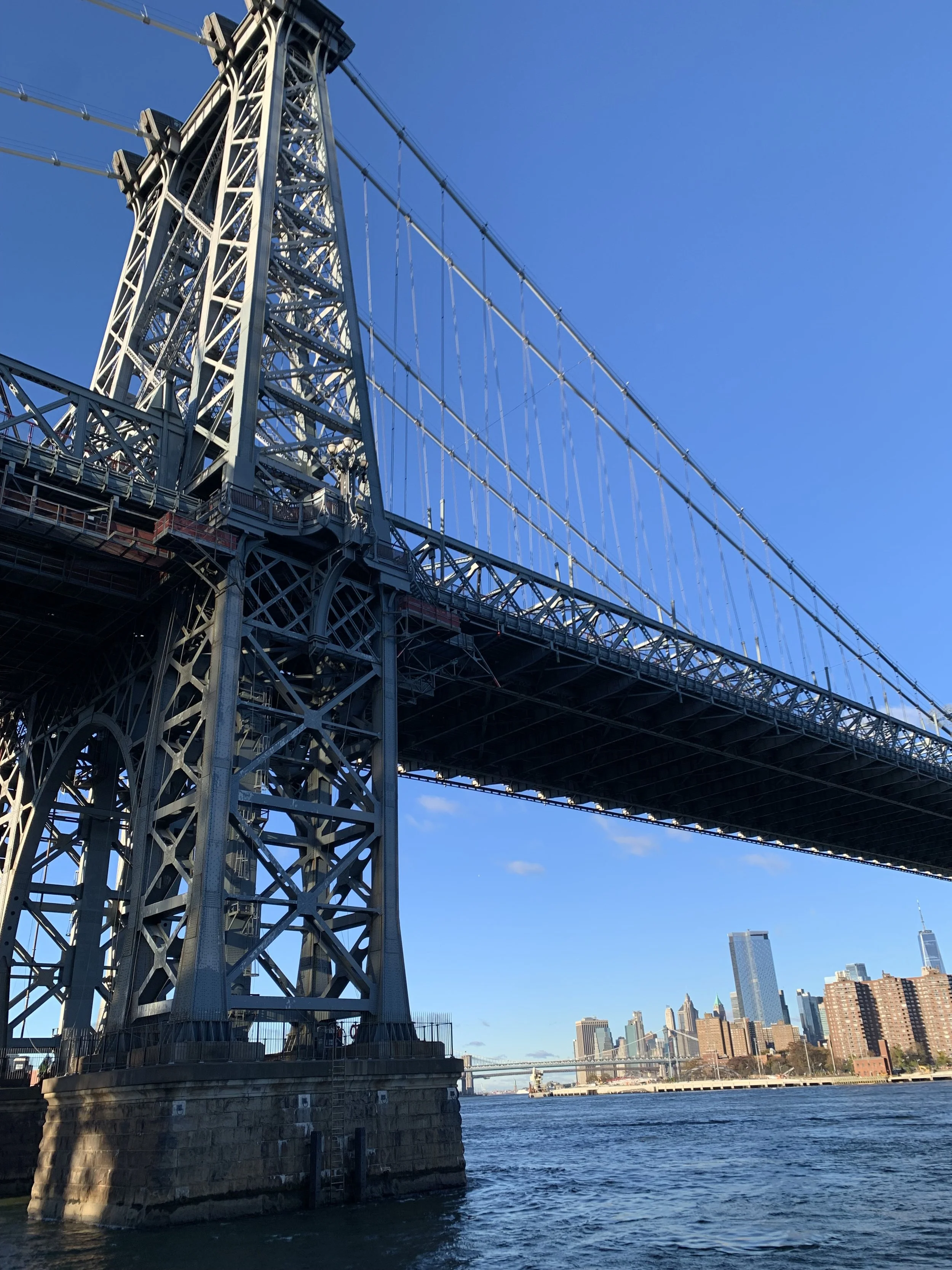View of the Manhattan Bridge under a clear blue sky with Manhattan skyline in the background, seen from the water.