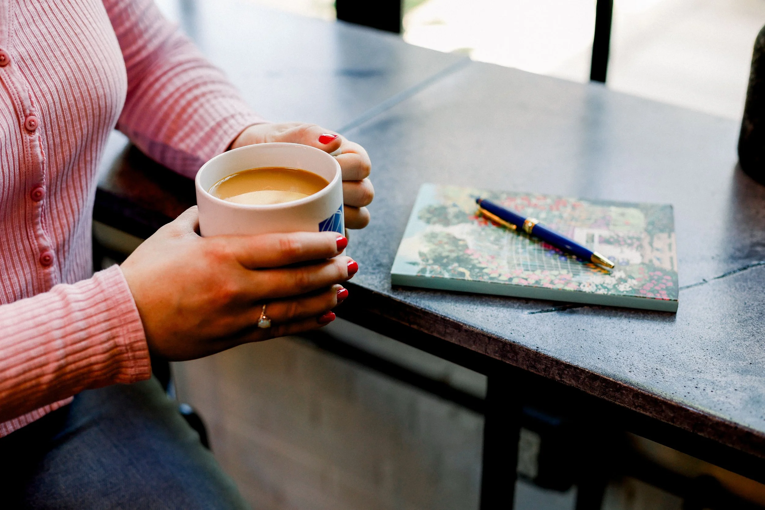 A person wearing a pink, ribbed shirt sitting at a table, holding a white mug filled with a hot beverage. There is a notebook with a colorful map cover and a blue pen on the table.