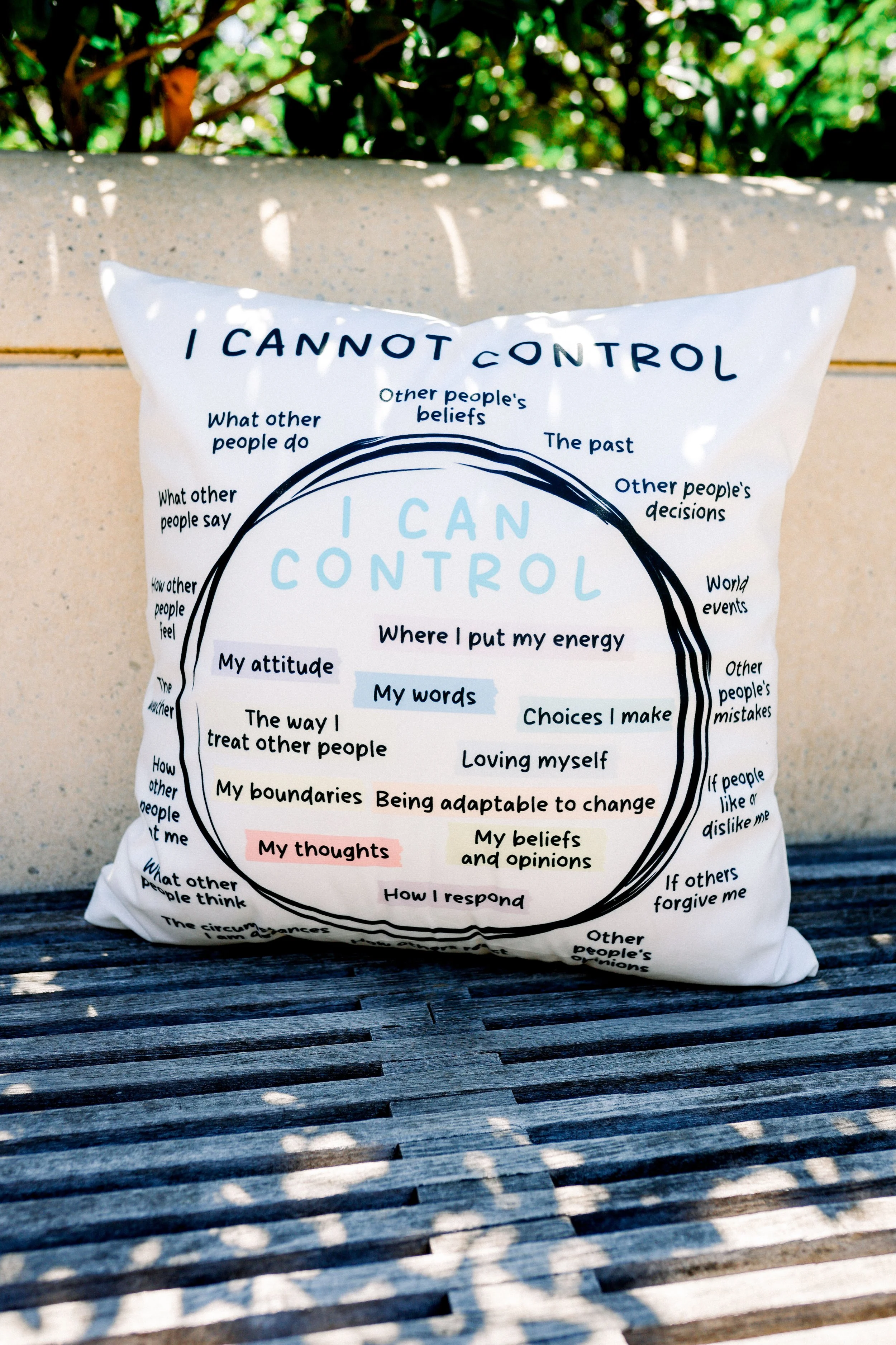 A white pillow with text about control and self-awareness, placed on a wooden surface outdoors with greenery in the background.