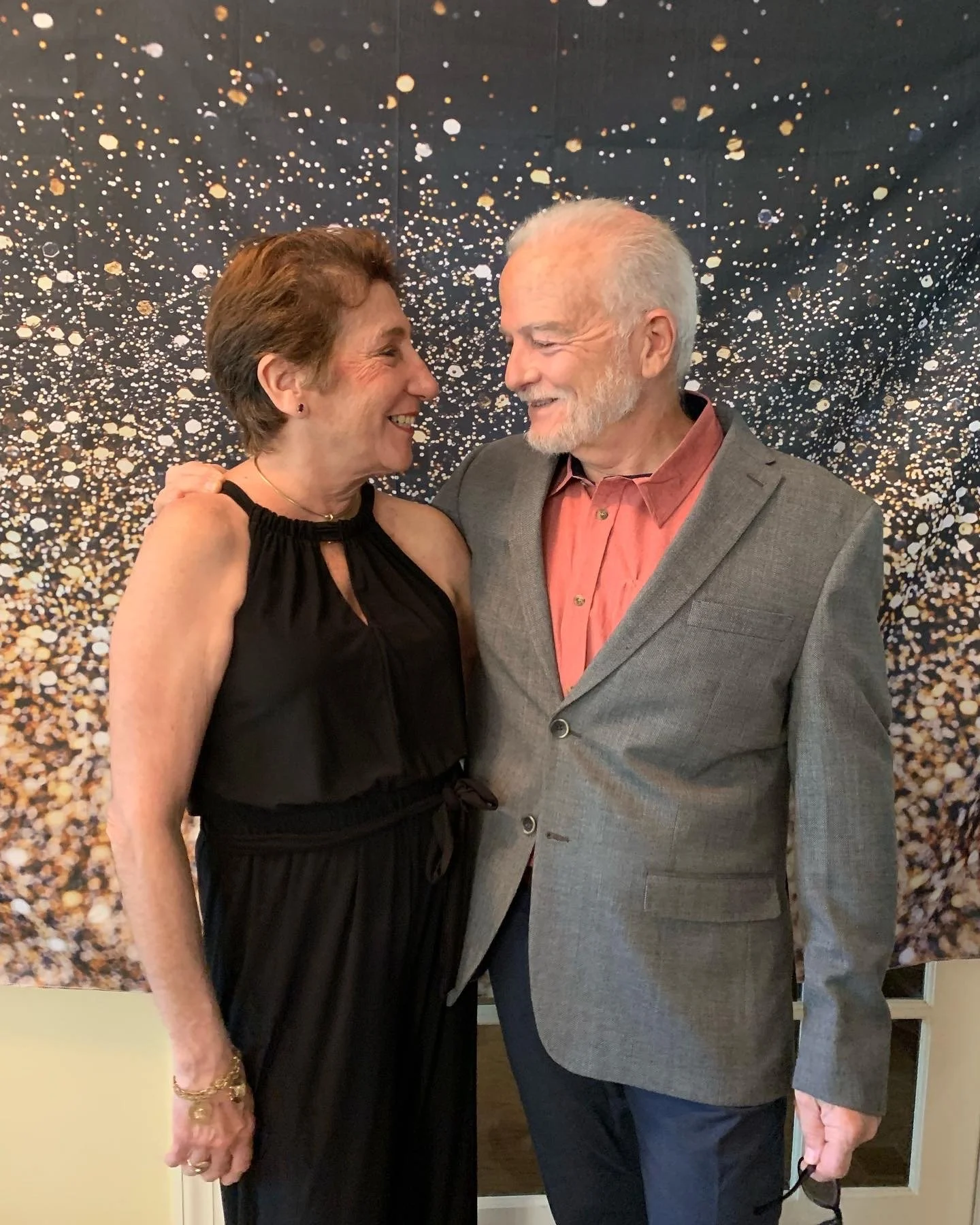 An elderly man and woman are standing close together, smiling and looking into each other's eyes. The woman is wearing a black dress with thin straps and gold jewelry. The man is wearing a gray blazer over a coral shirt and dark pants.