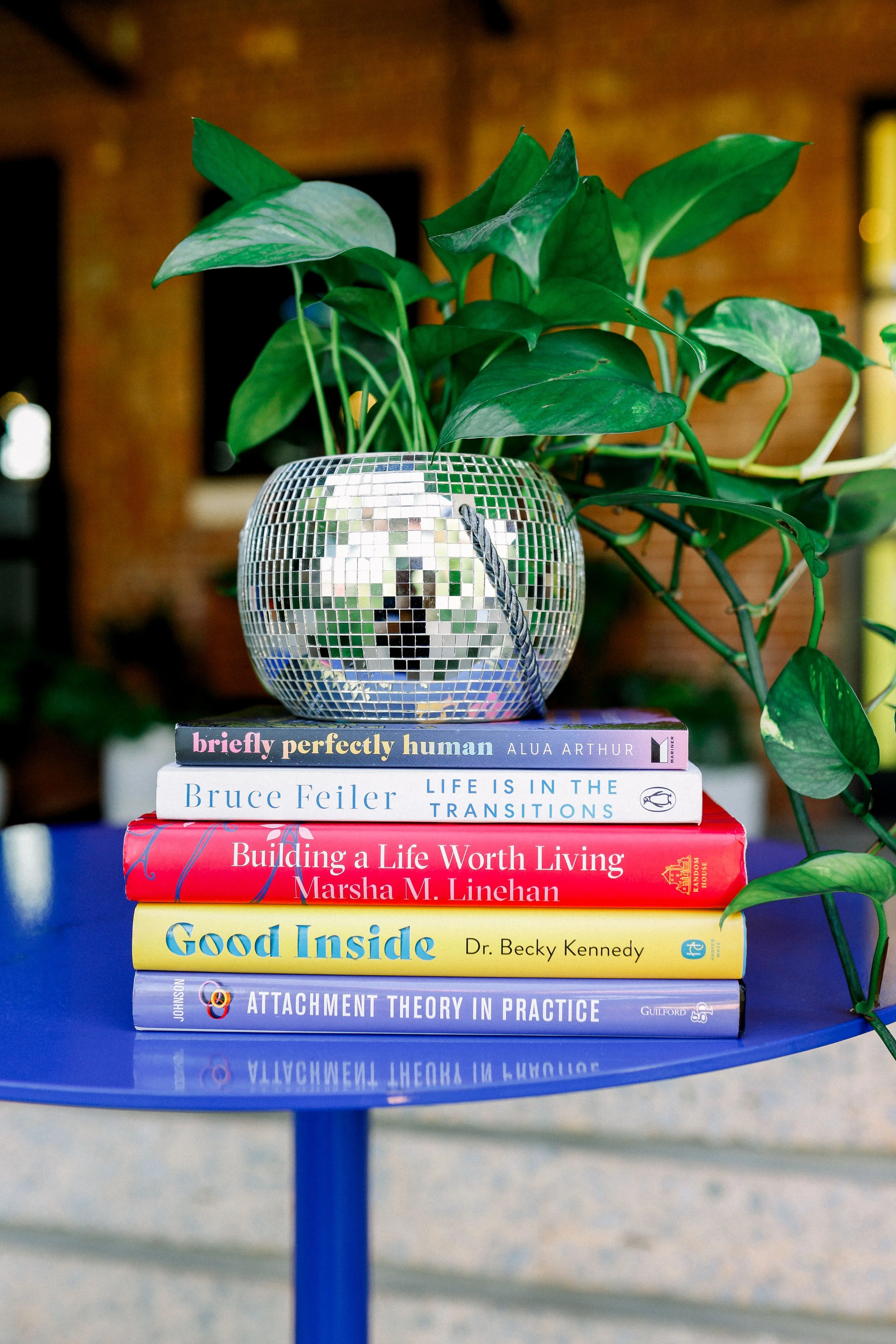 A stack of seven books on a blue round table, topped with a small, reflective mosaic globe-shaped sculpture. The background includes a brick wall and blurred window. The books focus on self-improvement, psychology, and personal growth.