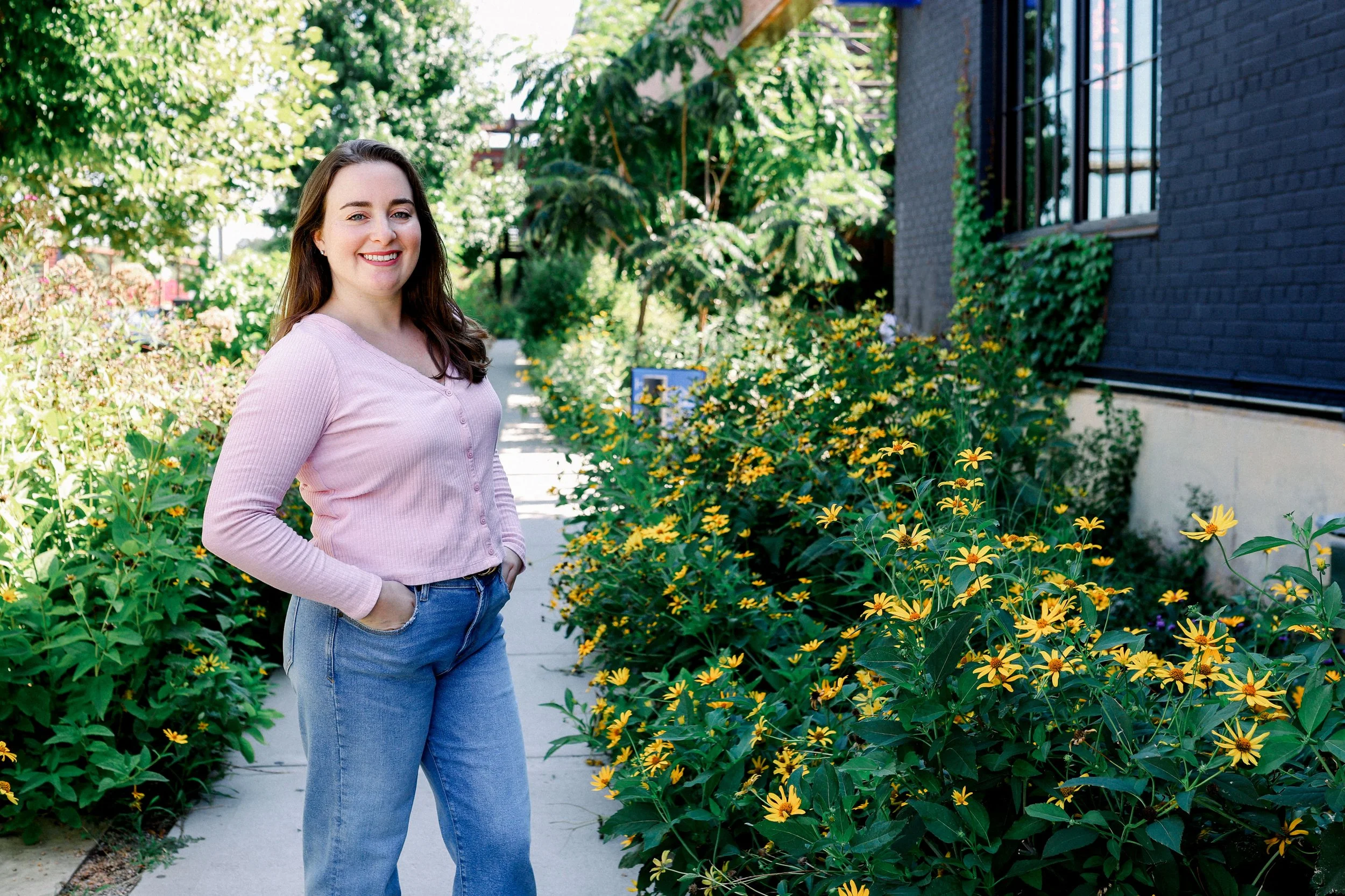 A woman with long brown hair, wearing a light pink long-sleeve shirt and blue jeans, stands on a sidewalk beside vibrant yellow flowers and lush green foliage in a garden area, smiling at the camera