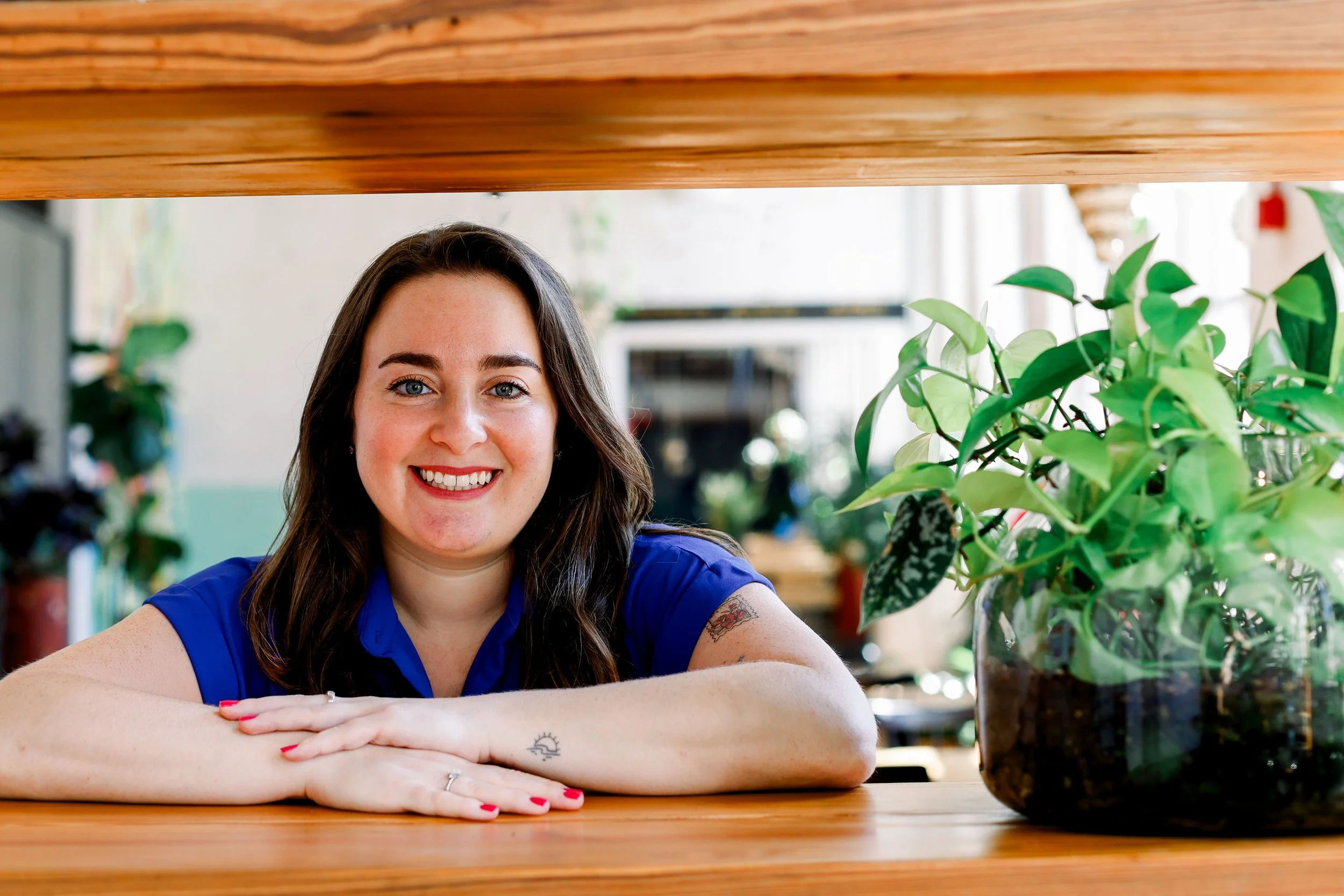A woman with long brown hair and blue eyes smiling with arms resting on a wooden table, seated in a cozy indoor space filled with green plants.