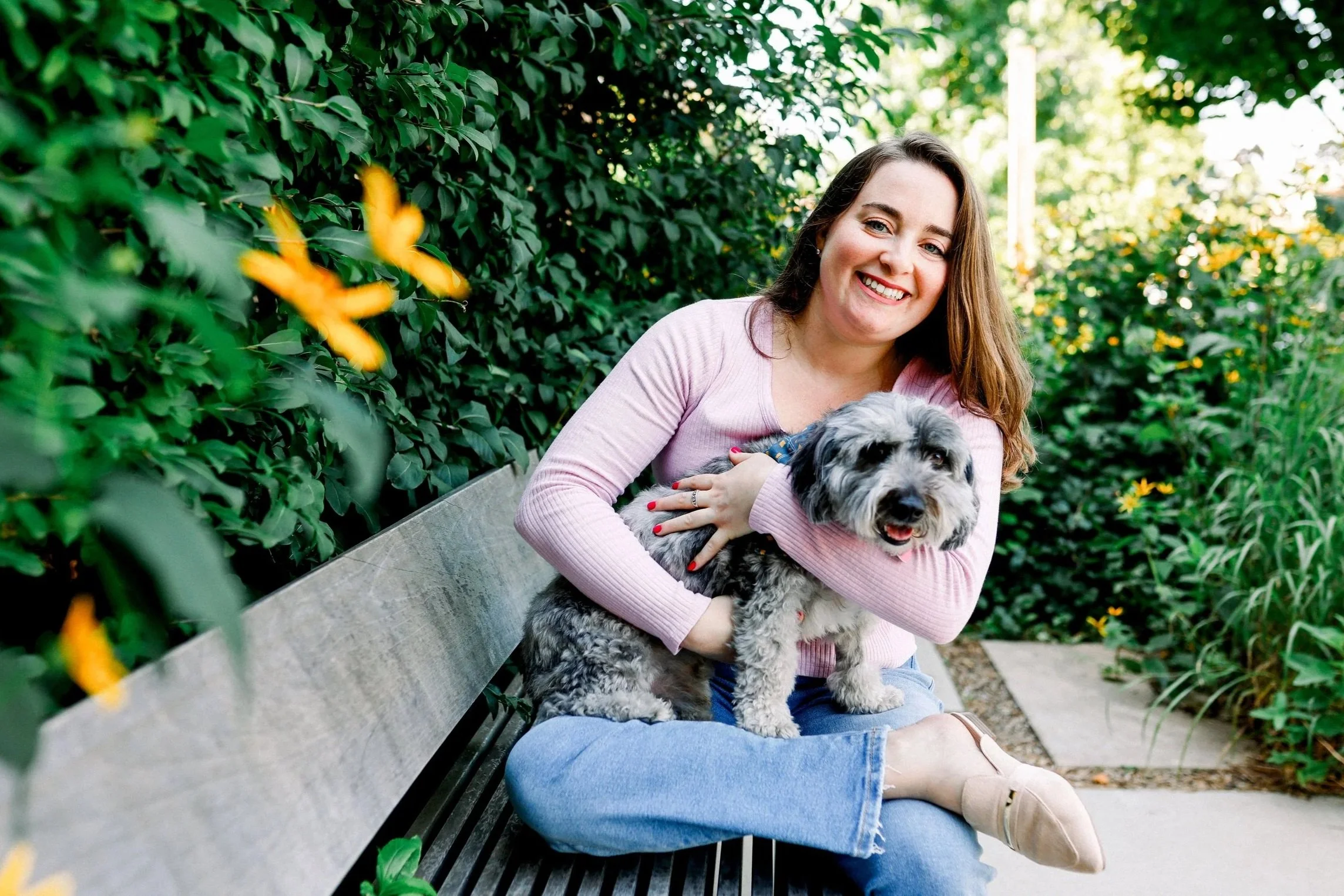 A woman with long brown hair, wearing a pink sweater and jeans, sitting on a park bench, smiling and holding a small, fluffy gray and black dog. There is greenery and yellow flowers around her.