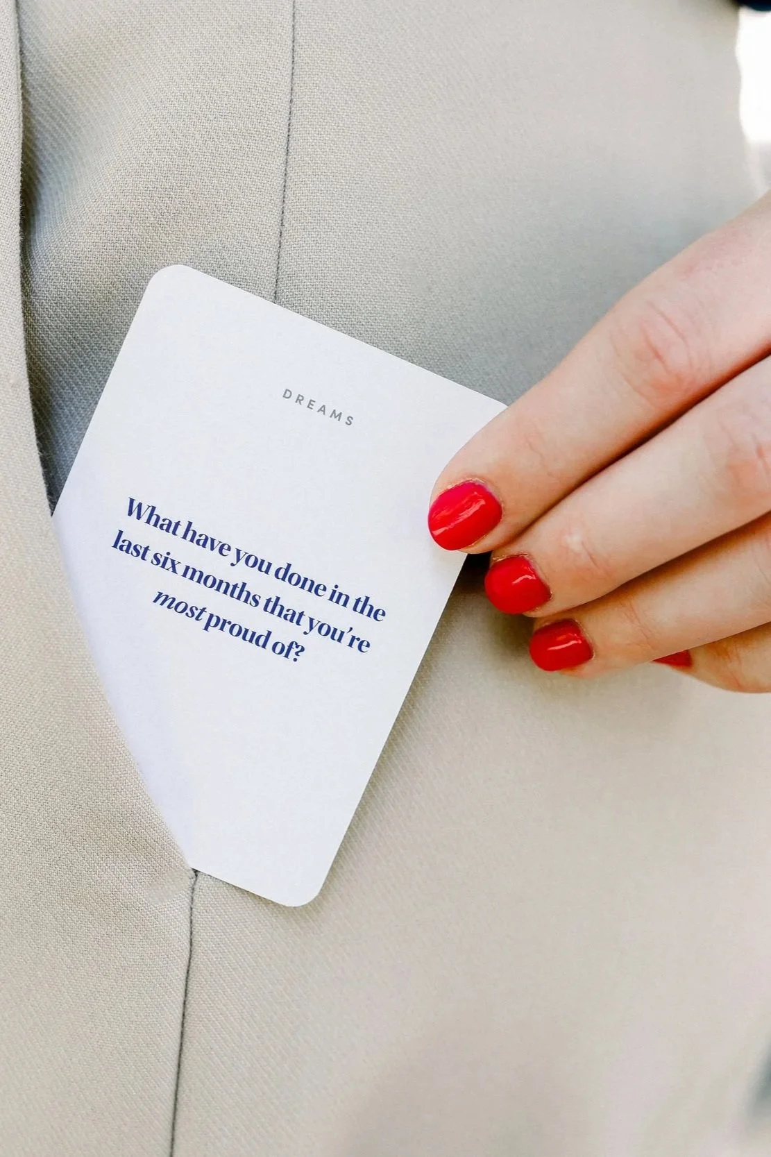 A person with red painted nails holding a white card that has the question, "What have you done in the last six months that you're most proud of?" printed on it, against a beige fabric background.