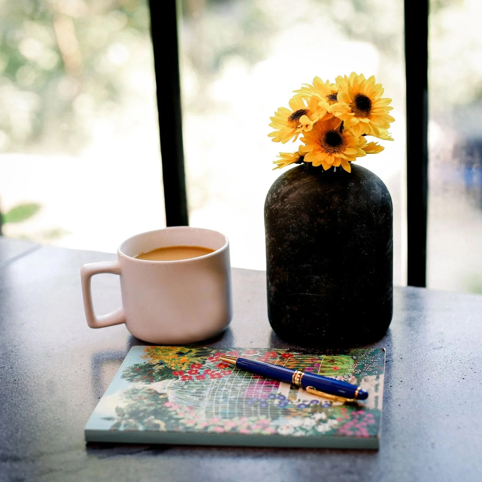 A white mug filled with coffee, a colorful puzzle with a blue pen on top, and a dark vase with yellow flowers sitting on a table near a window.