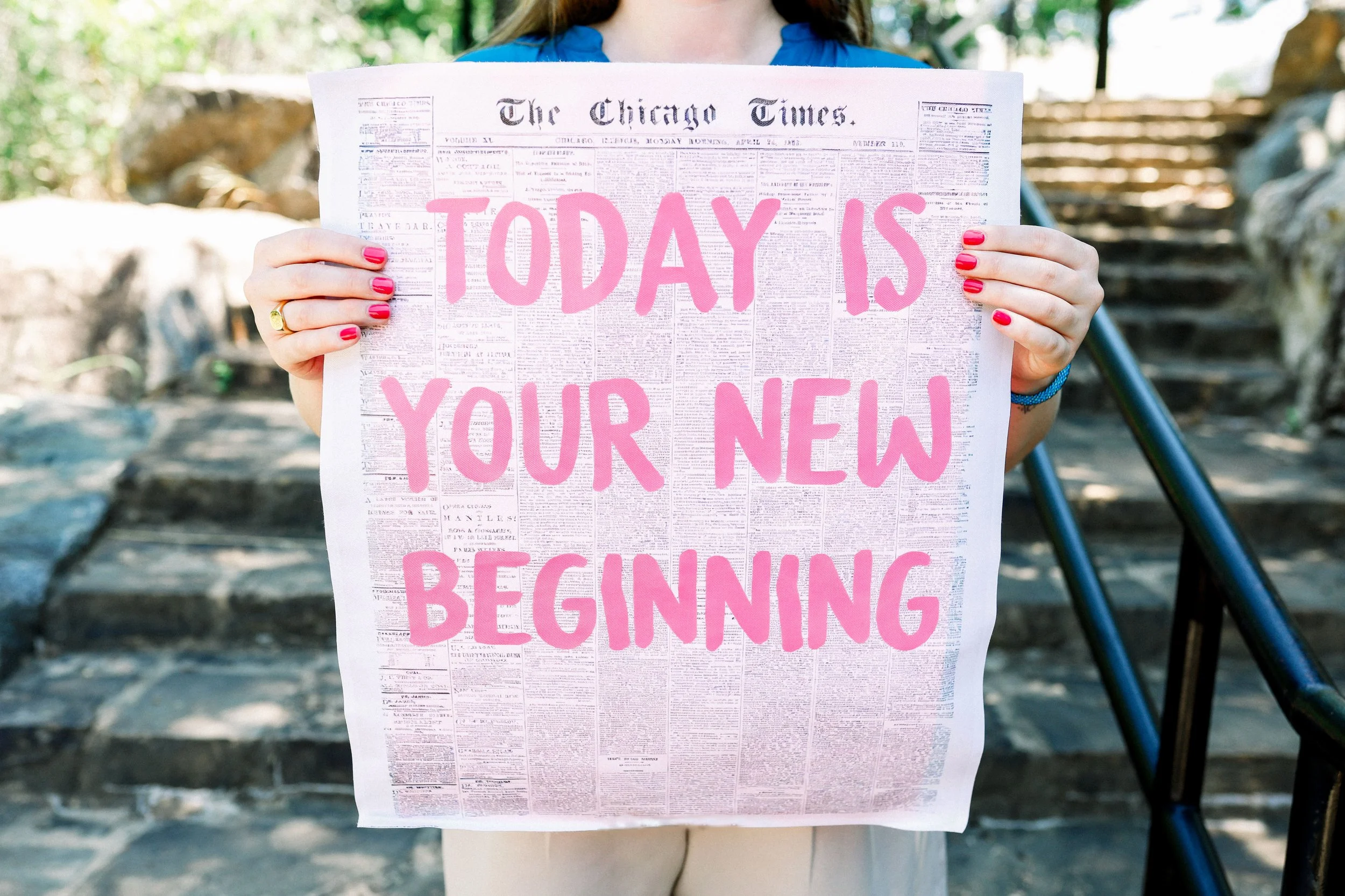 Person holding a newspaper with pink text that reads 'Today is your new beginning' at an outdoor location with stone steps and greenery in the background.