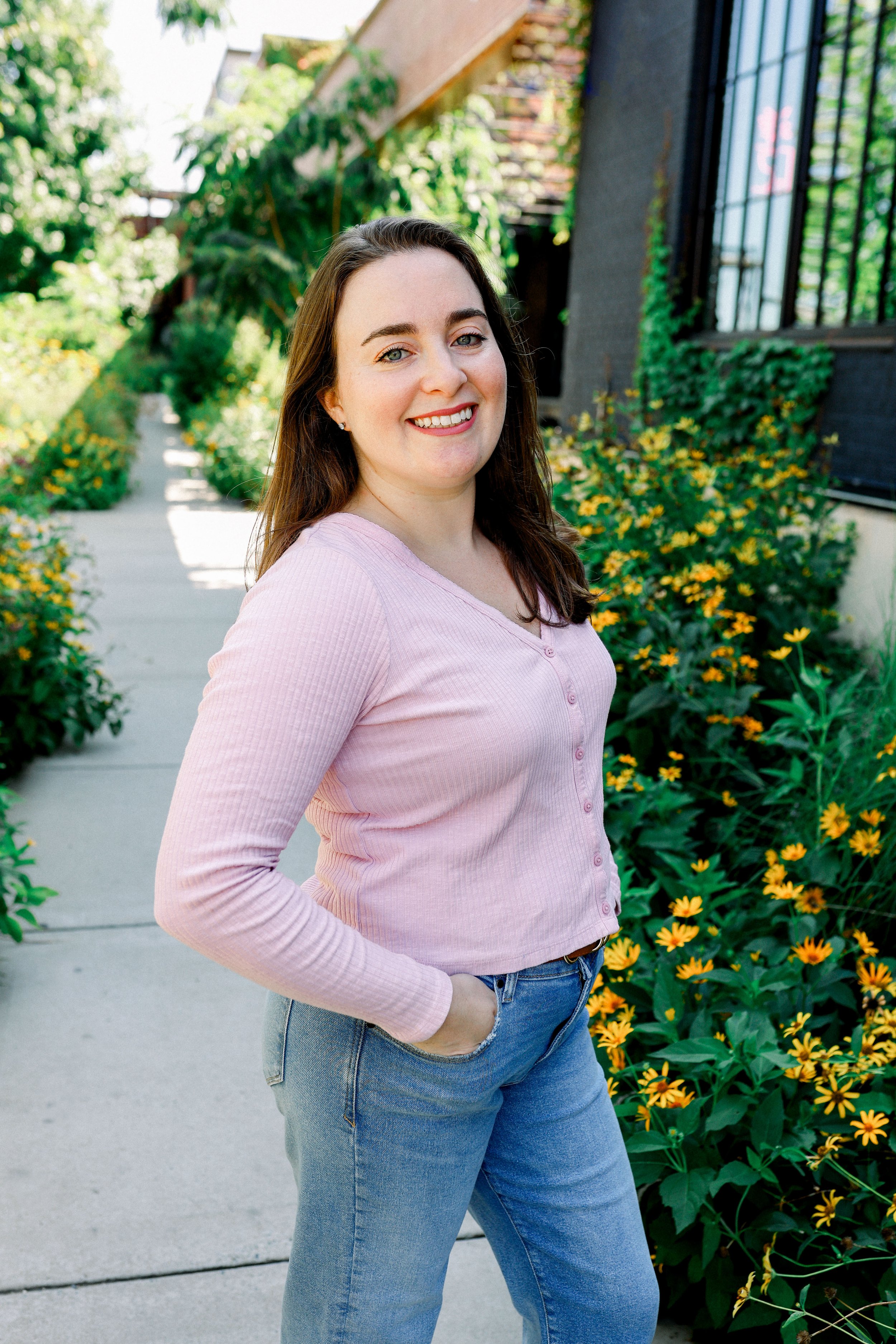 A smiling woman with brown hair, wearing a pink shirt and blue jeans, standing on a sidewalk next to a row of yellow flowers and green bushes in a residential area.